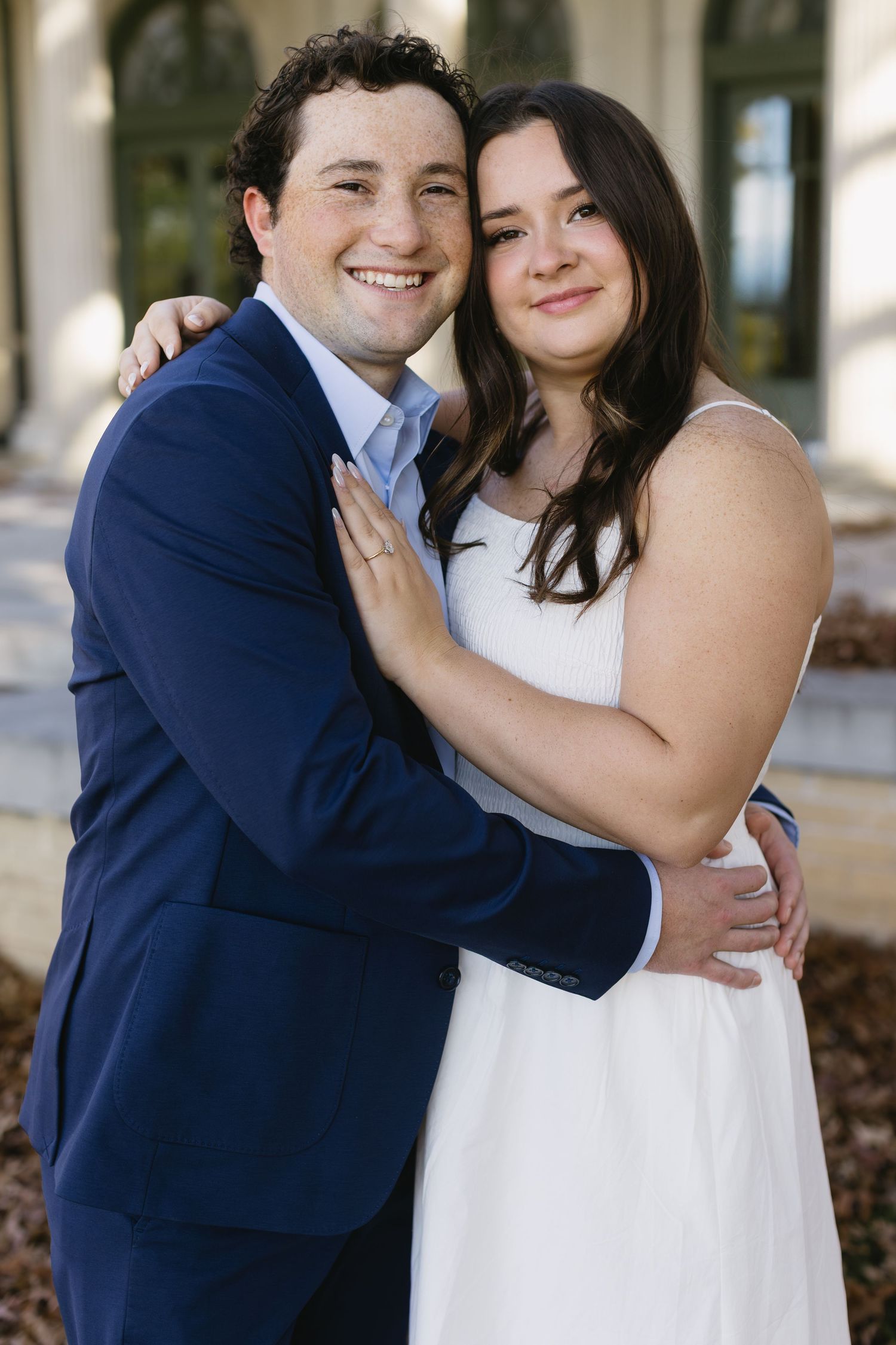 A happy couple poses together outdoors, with one wearing a navy suit and the other in a white dress.