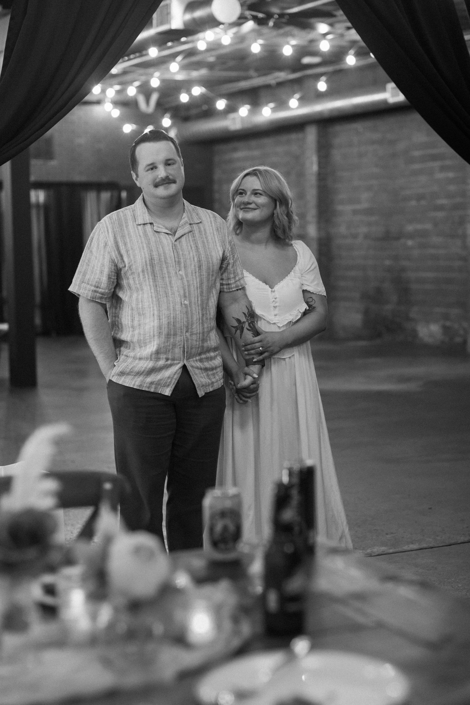 A vintage-style black and white photo showing a couple at an indoor venue with string lights.