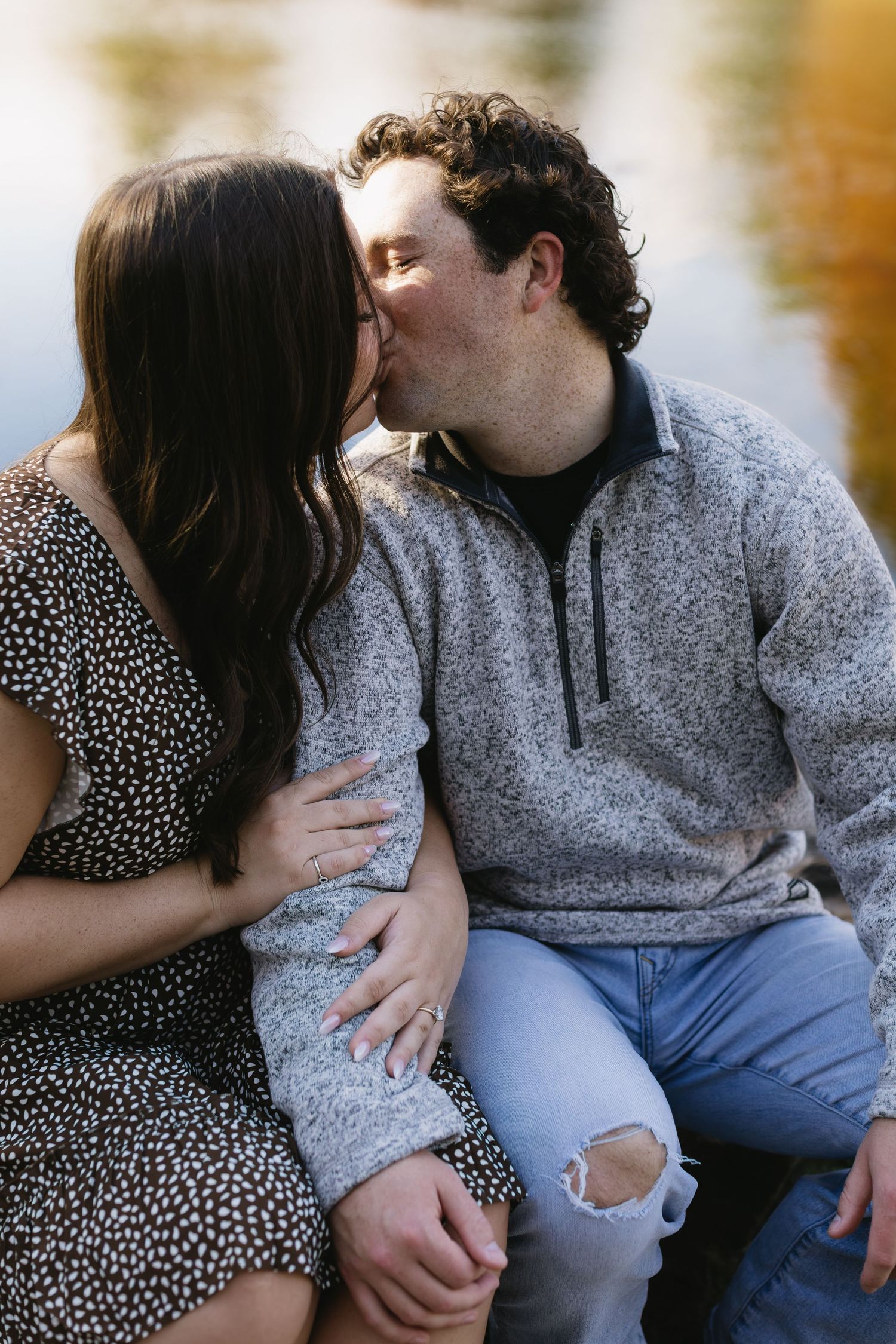 A couple sits close together by a lake wearing casual clothing during an engagement session.