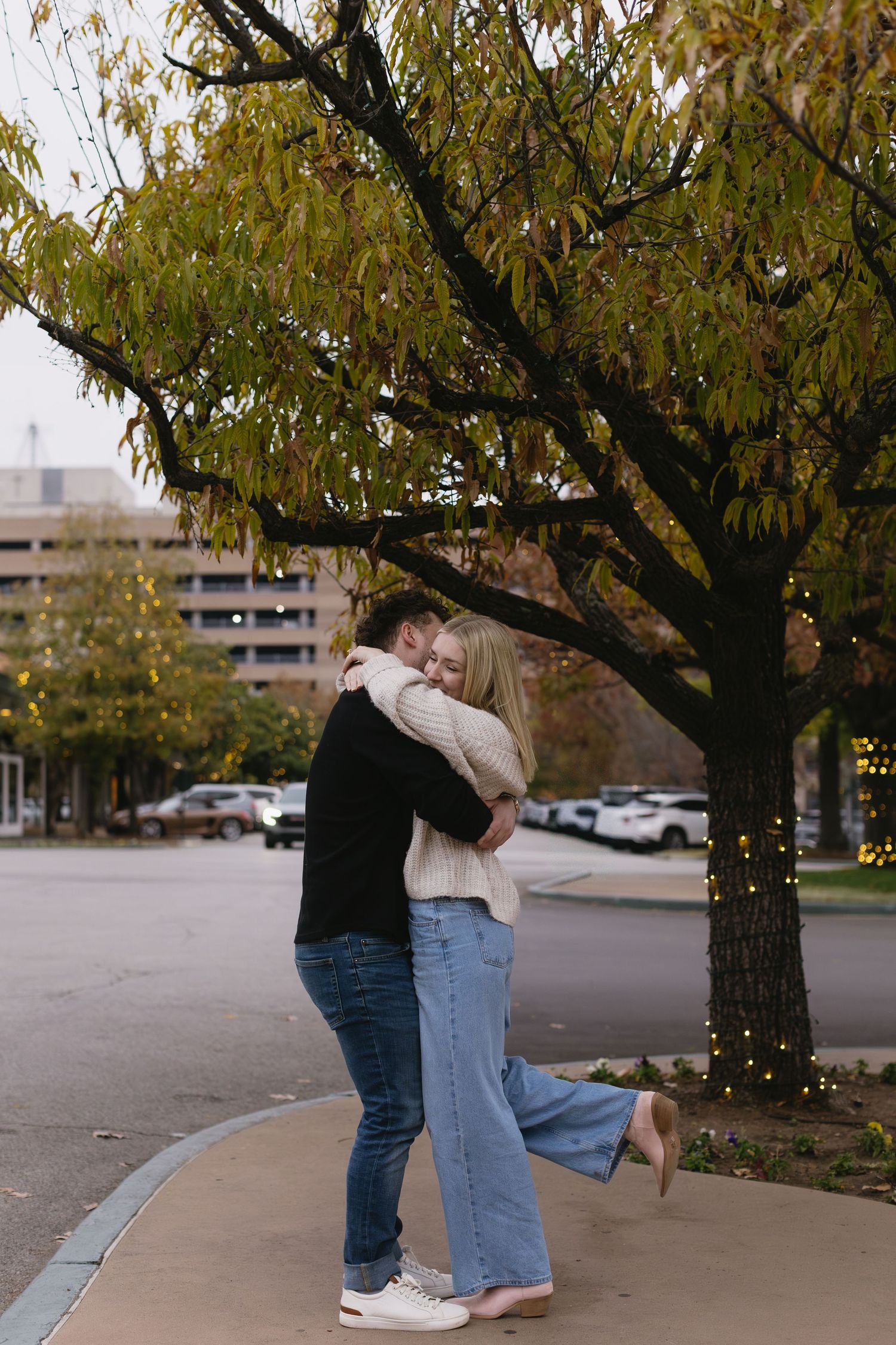 A couple shares tender moments under a tree with fall leaves in an urban setting.