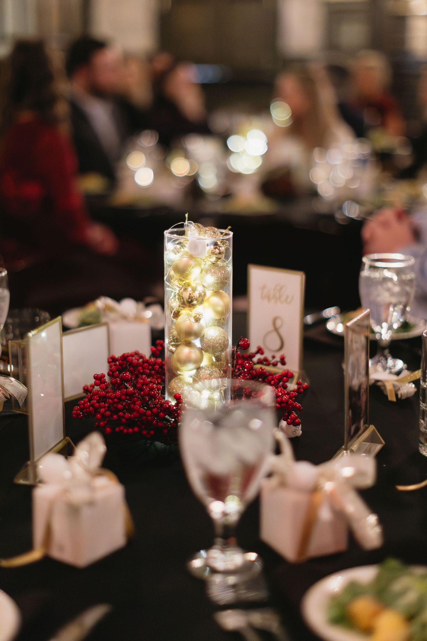 Elegant wedding reception table with red berries, fairy lights in mason jars, and place cards on black tablecloth.