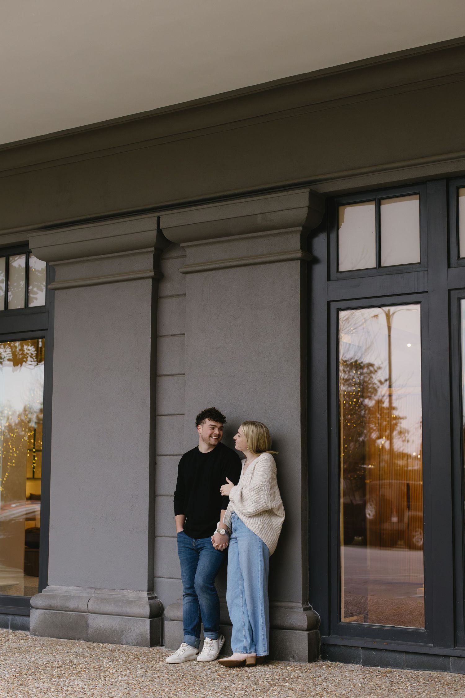 A couple poses together against a gray building exterior with large windows.