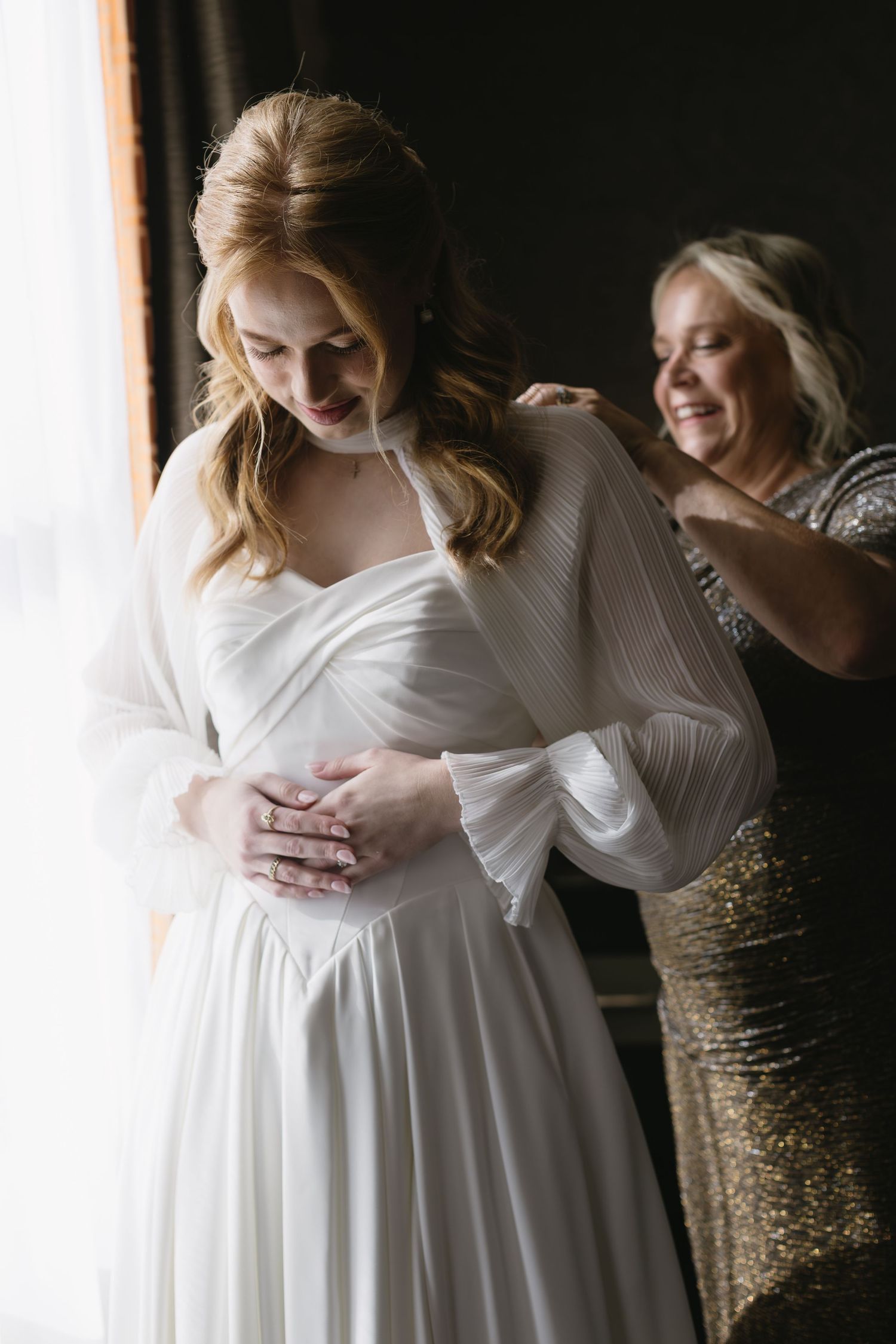 A bride in a flowing white gown with long sleeves stands in soft window light.