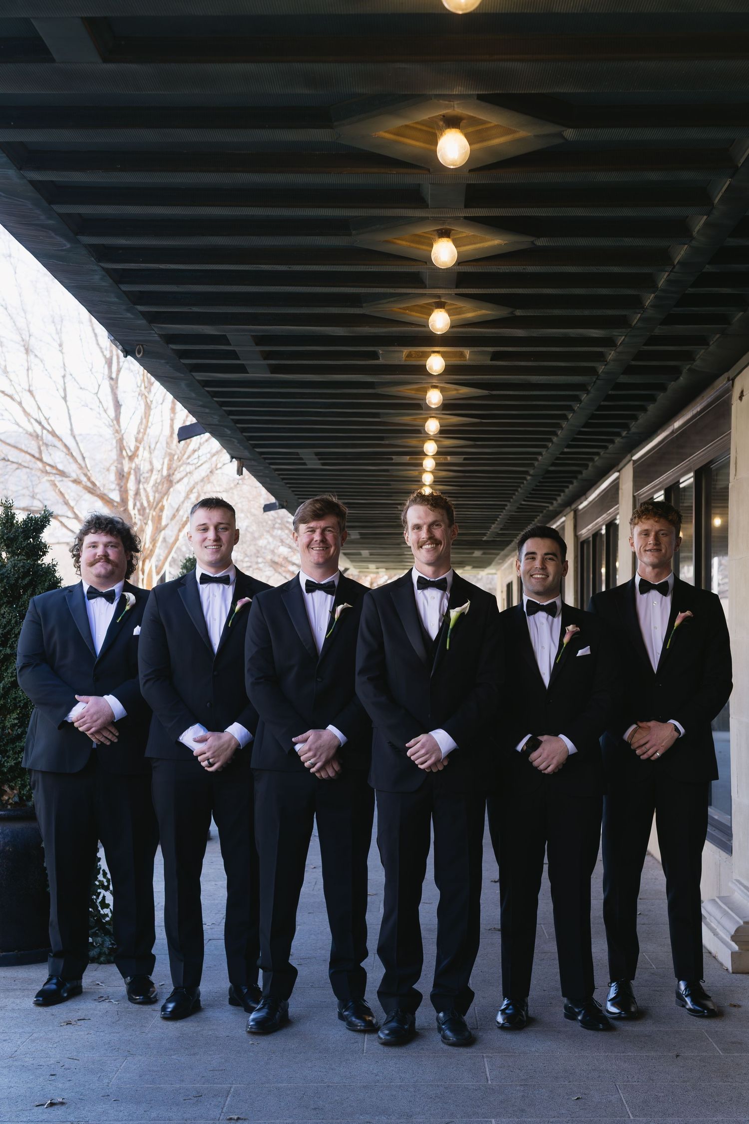 Groomsmen lined up in black tuxedos under covered walkway.