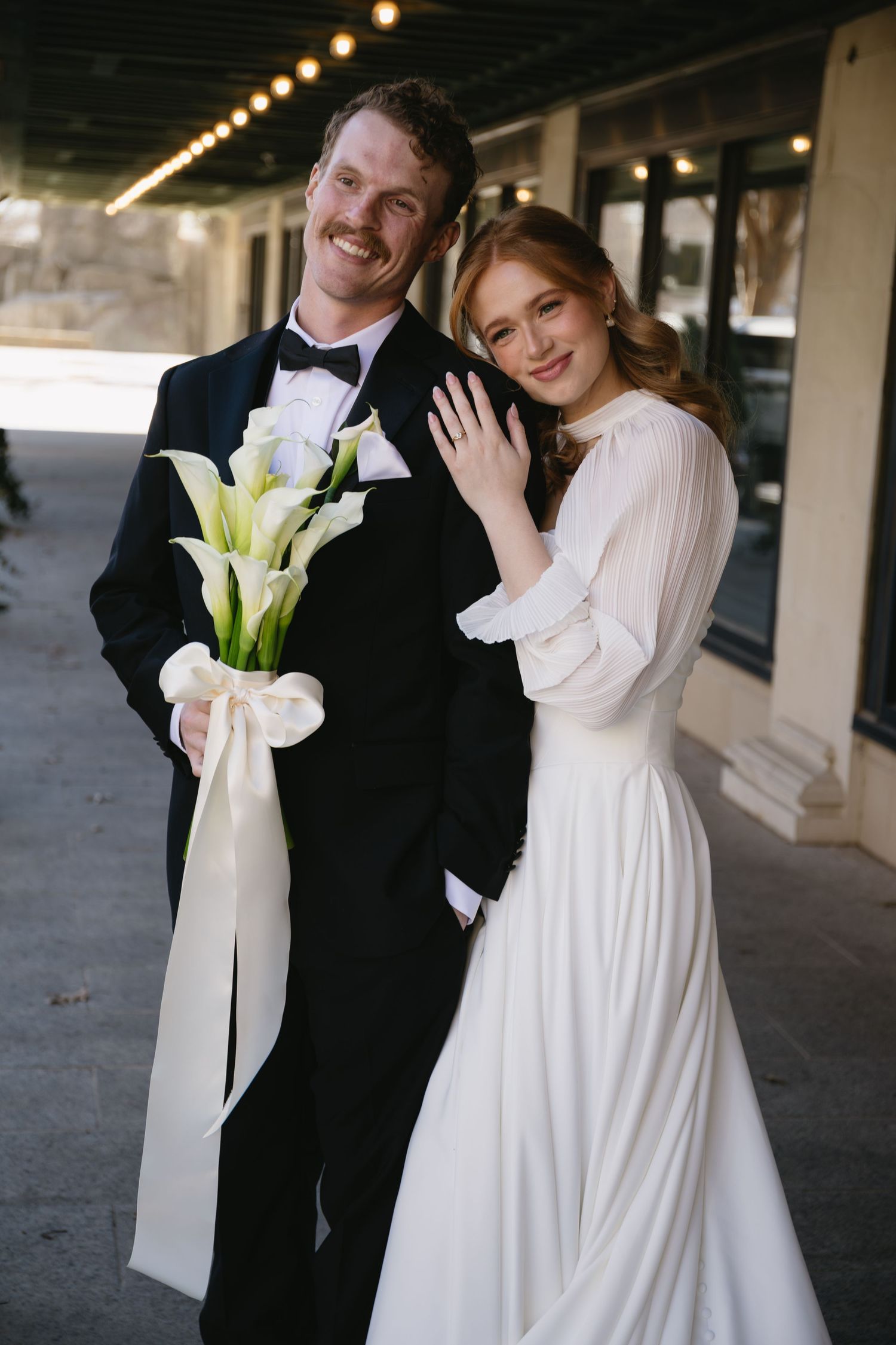 Wedding couple poses together outdoors with calla lily bouquets and classic black and white attire.
