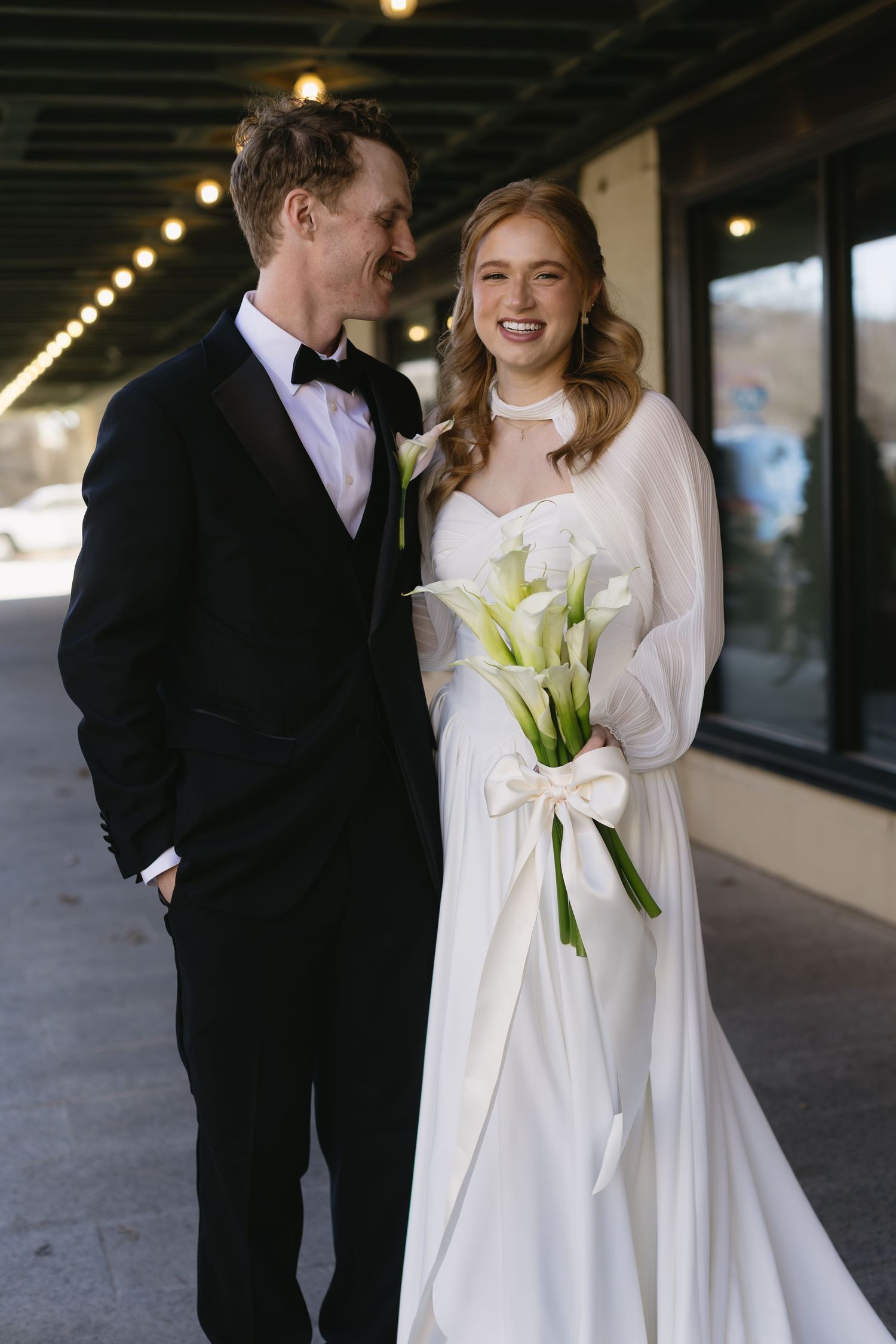 A couple shares a joyful moment outside a building, with the bride holding elegant white calla lilies.