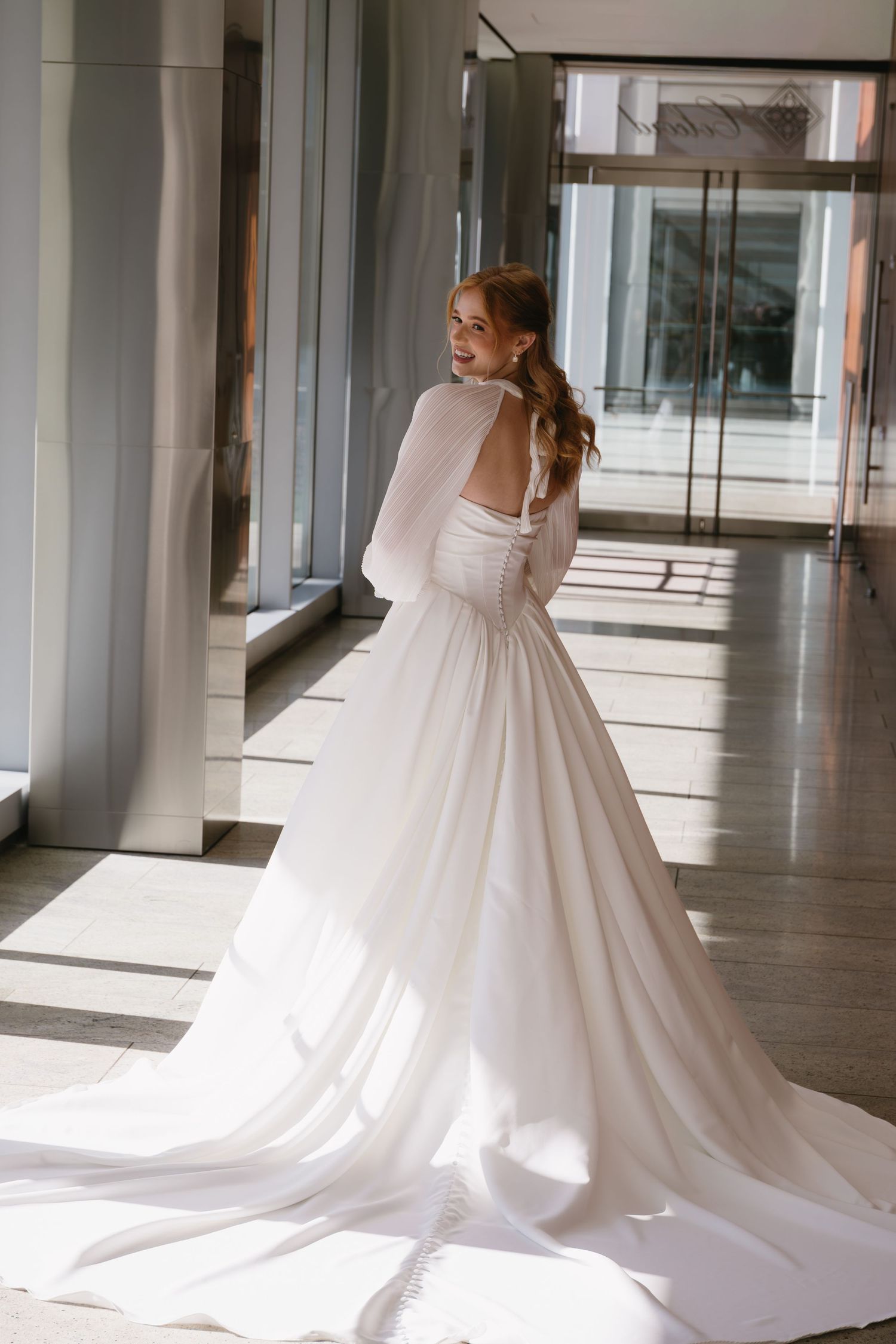 Elegant wedding gown with flowing train photographed in modern hallway with natural lighting.