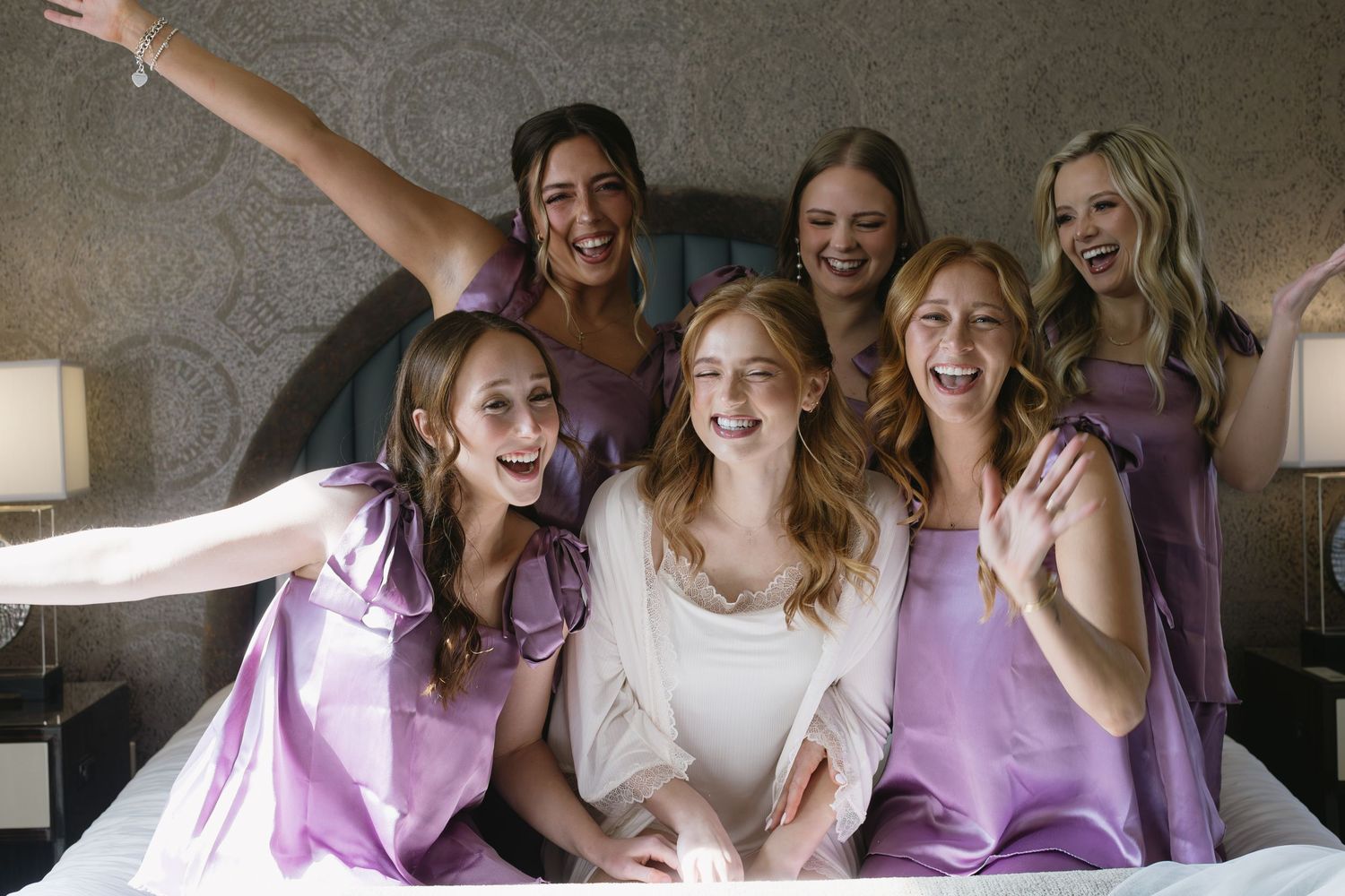 Wedding party wearing coordinated lavender and white dresses posing together on a bed.