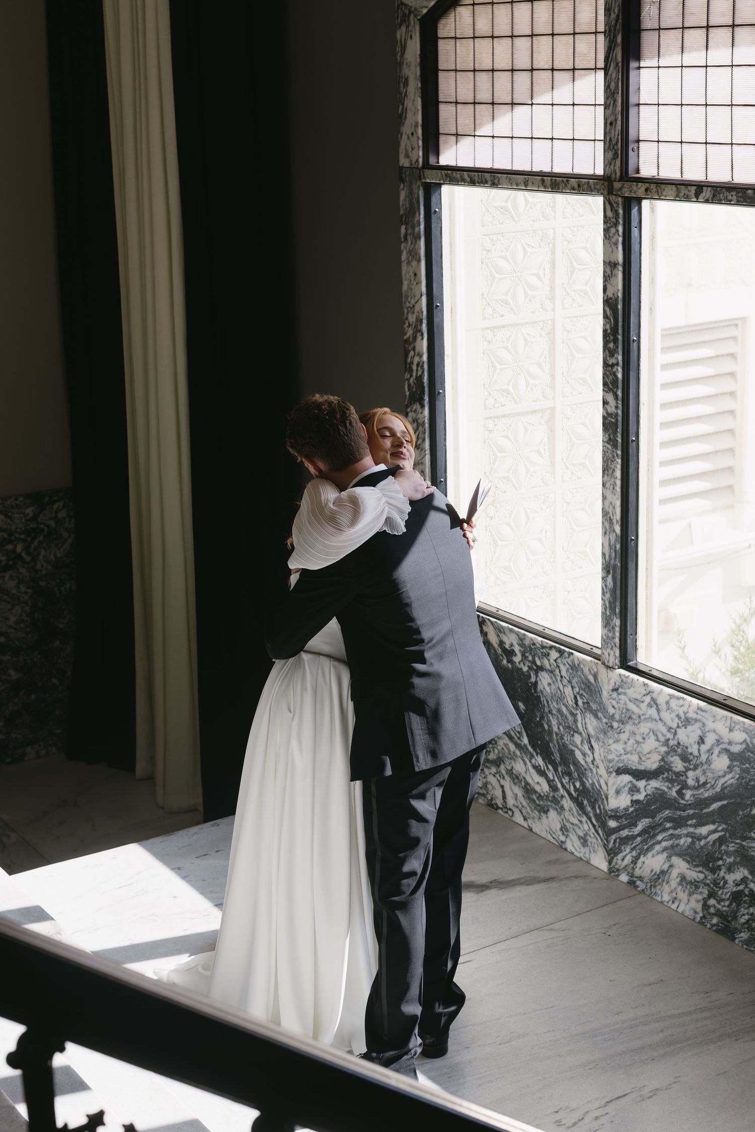 A couple embraces in an elegant indoor setting with marble walls and large windows.