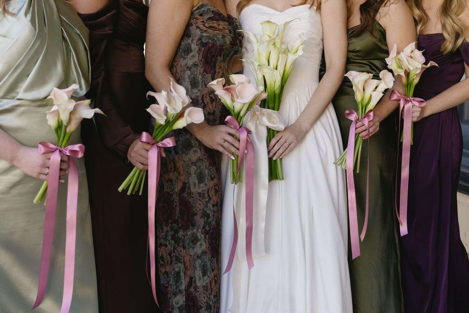 Bridesmaids holding elegant calla lily bouquets with pink ribbons at a wedding ceremony.