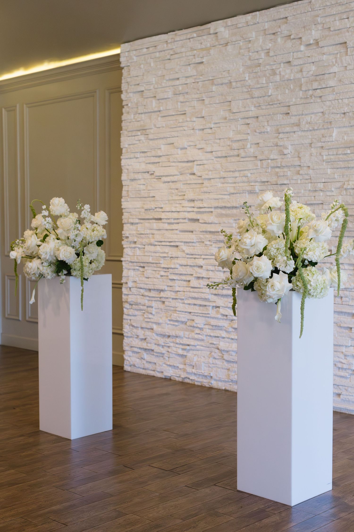 Modern white floral arrangements in geometric white pedestals against a textured stone wall.