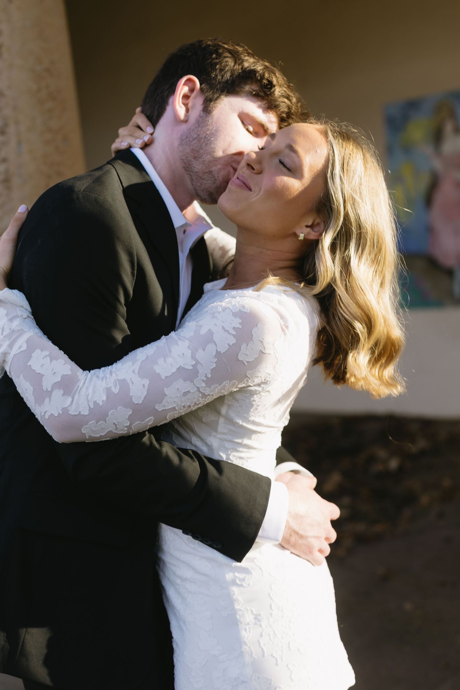 A romantic wedding photo of a couple embracing, with the bride in a long-sleeved lace dress and groom in black suit.
