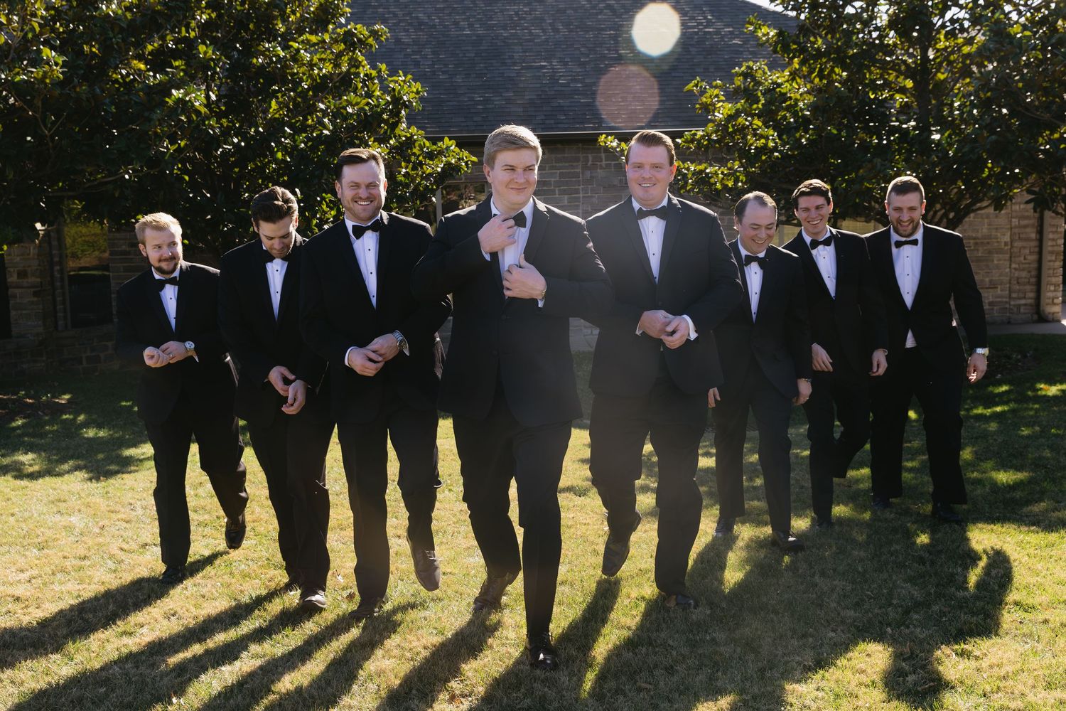 Groomsmen in black tuxedos pose together outdoors on sunny day.