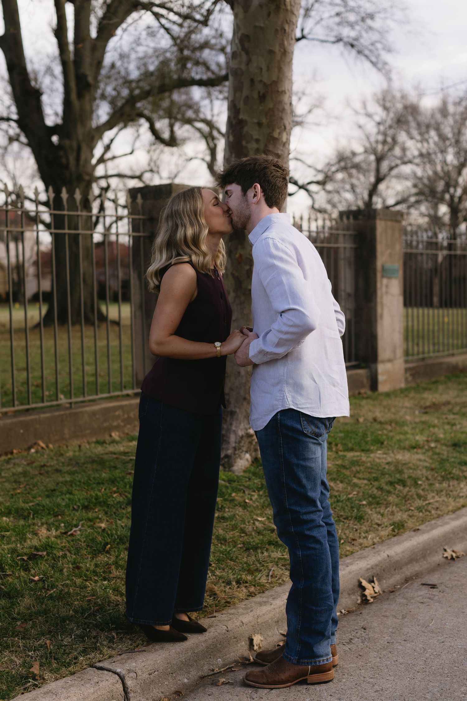 A casual couple sharing intimate moments along a tree-lined path with iron fencing.
