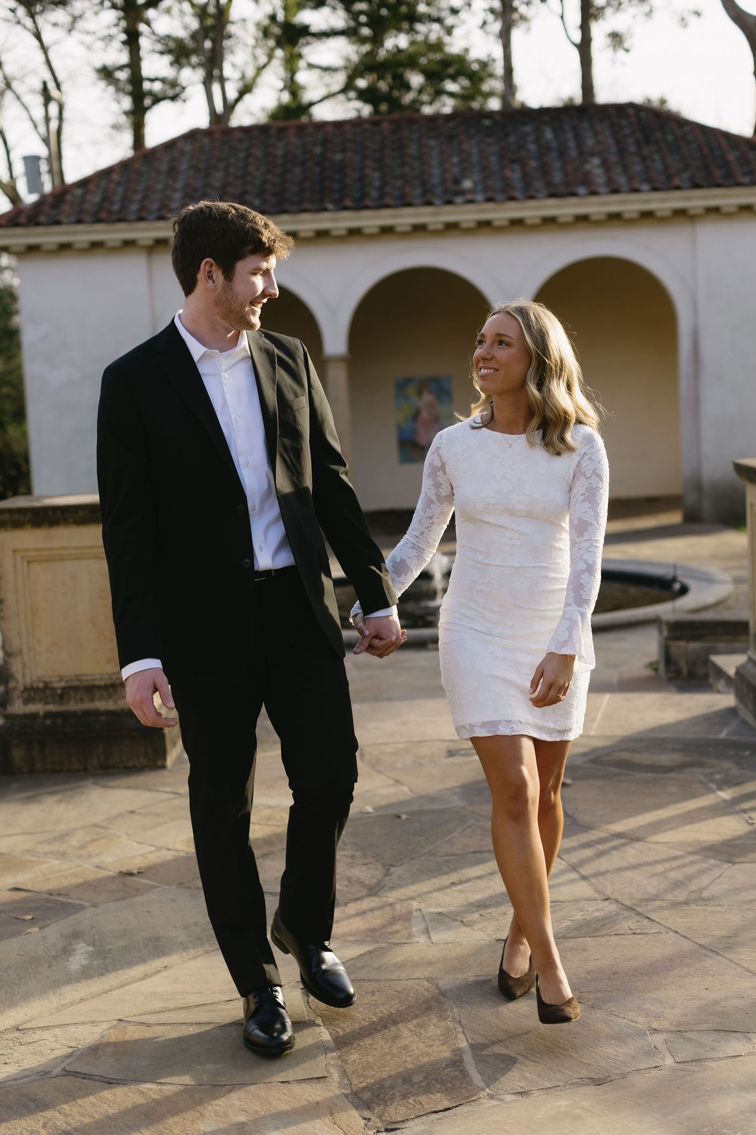 A couple in formal attire walks together on a stone patio with arched architecture in the background.