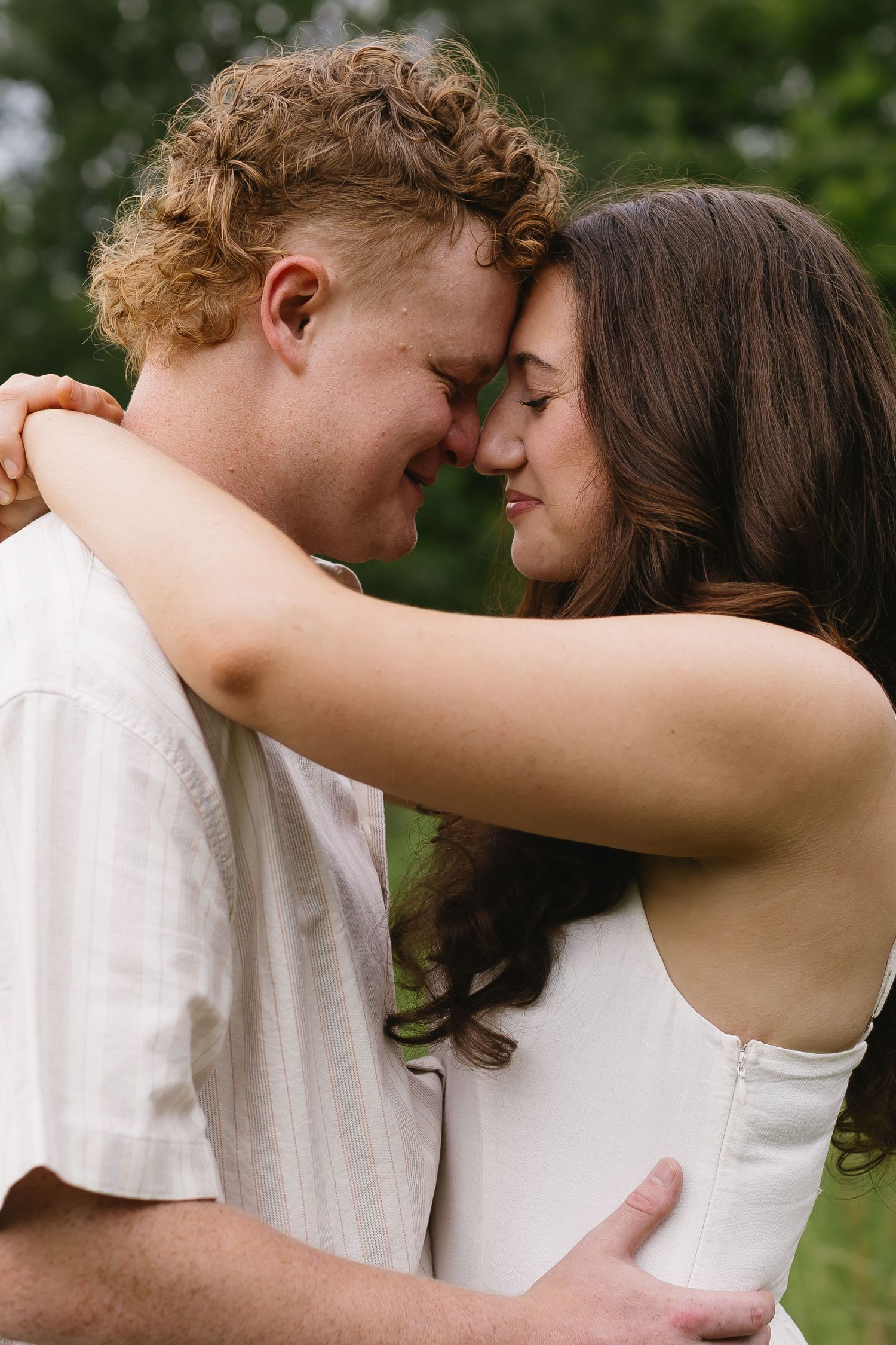 A romantic couple embraces in a green summer field wearing coordinating white outfits.