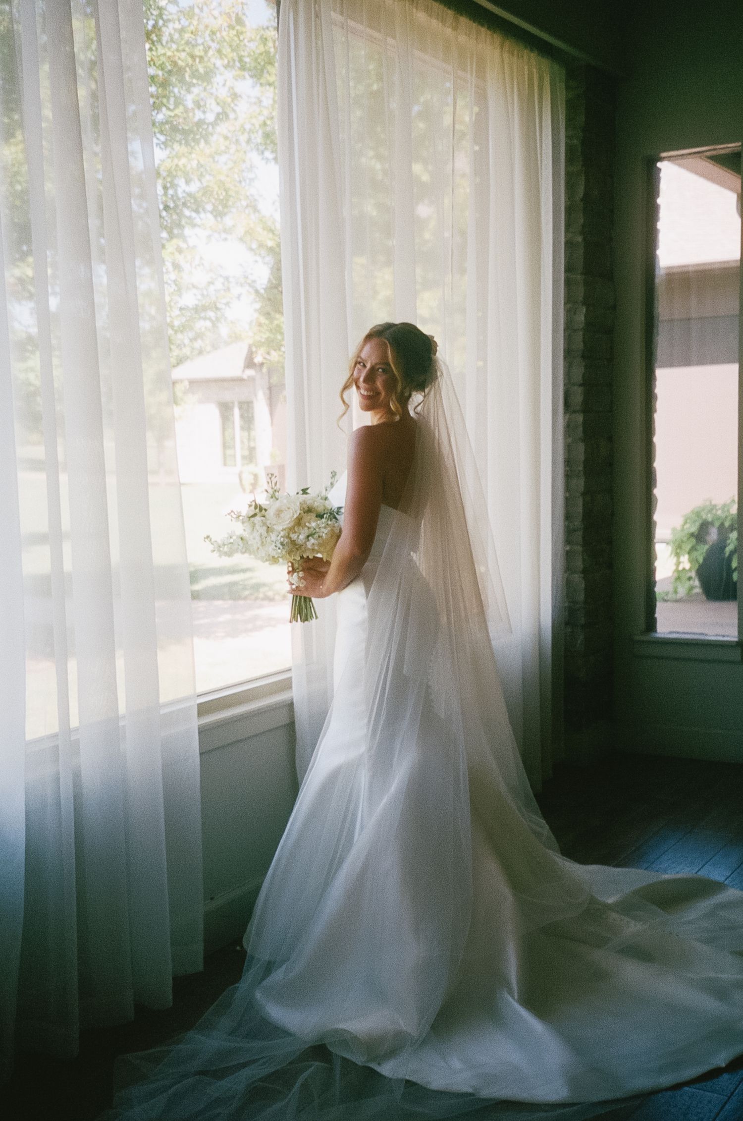 Bride in flowing white wedding dress poses near sheer curtained windows in a moody, atmospheric setting.
