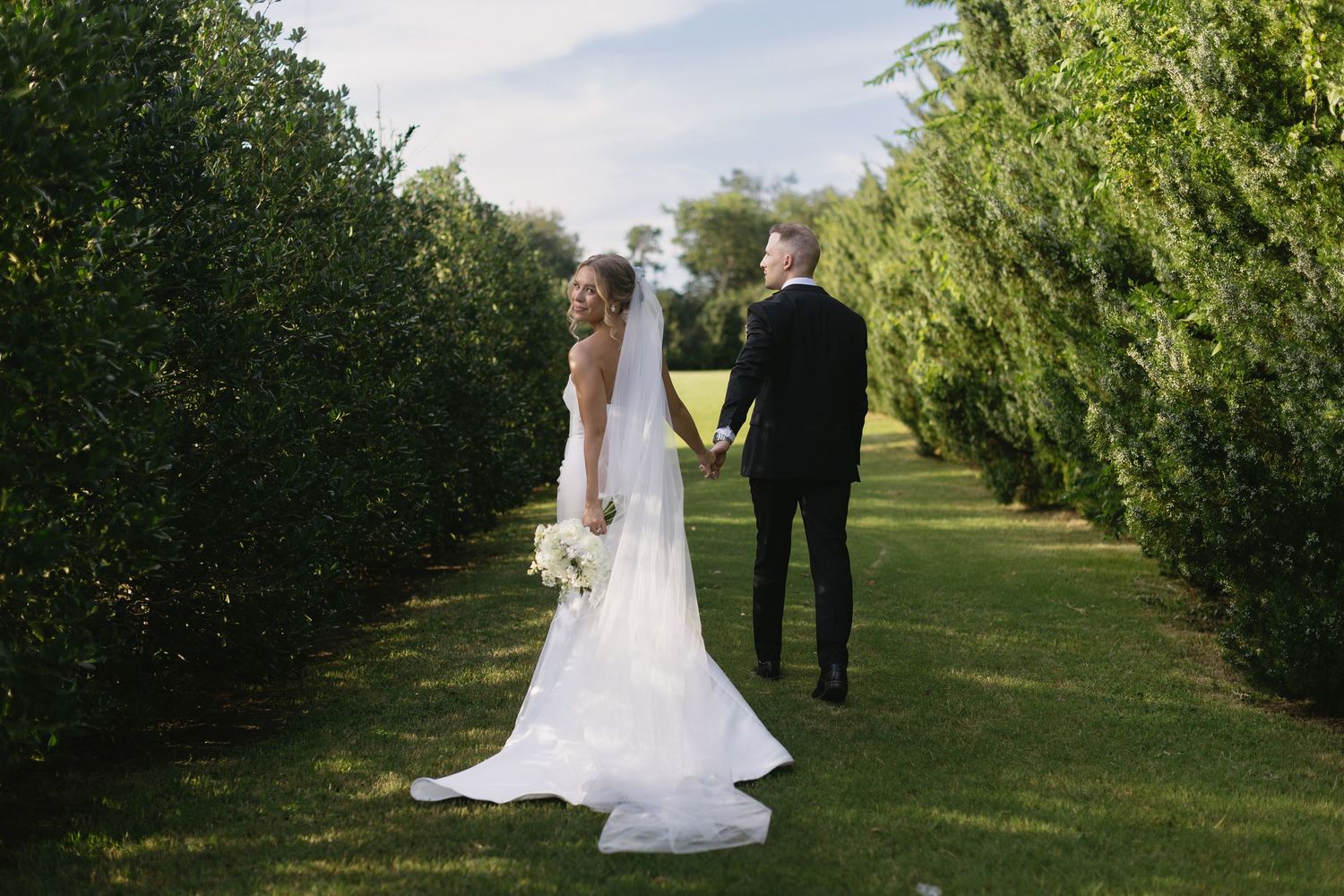 A wedding couple stands between rows of lush green trees in an orchard during a summer day.