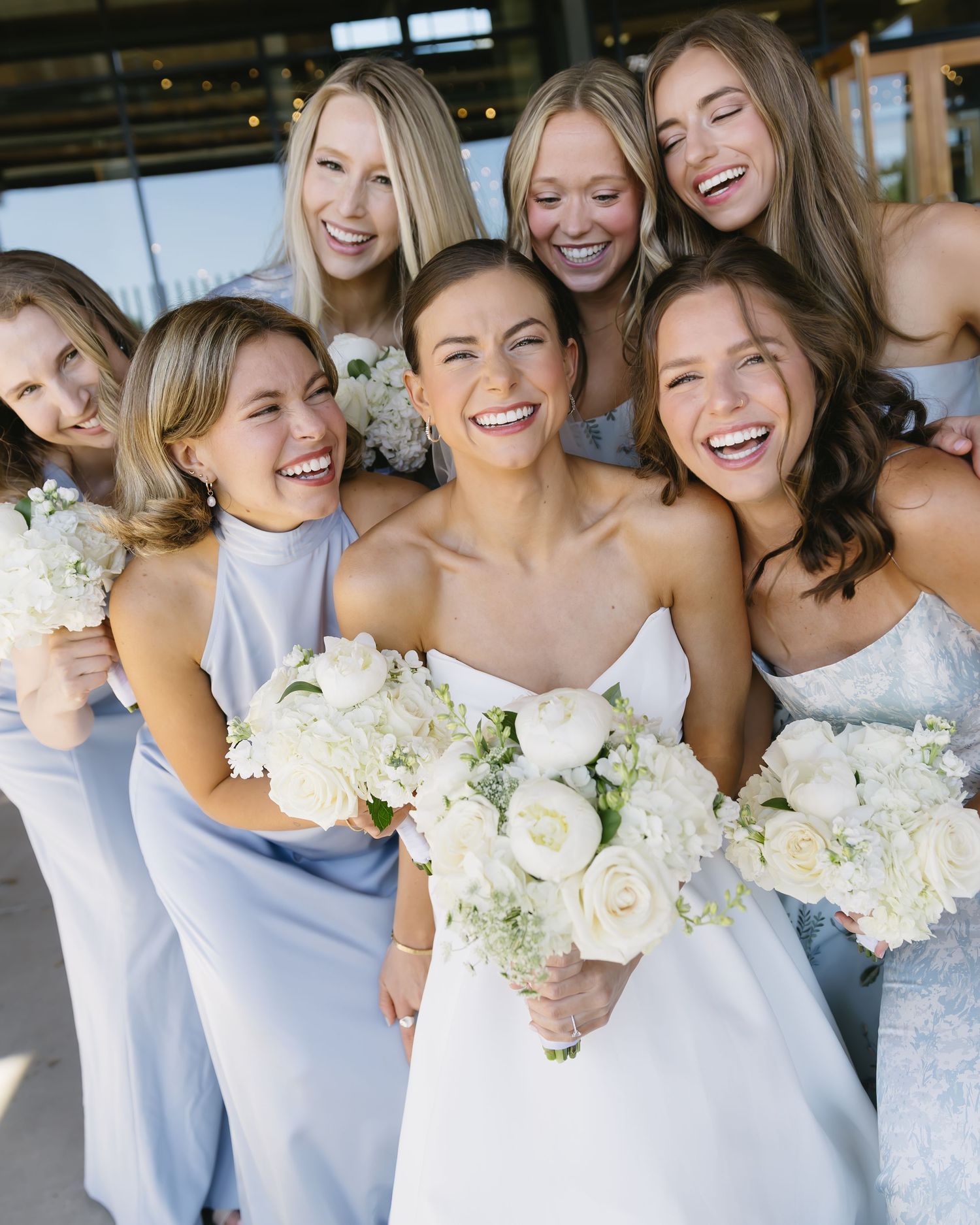 Bridal party in light blue dresses holding white bouquets share joyful moment together.
