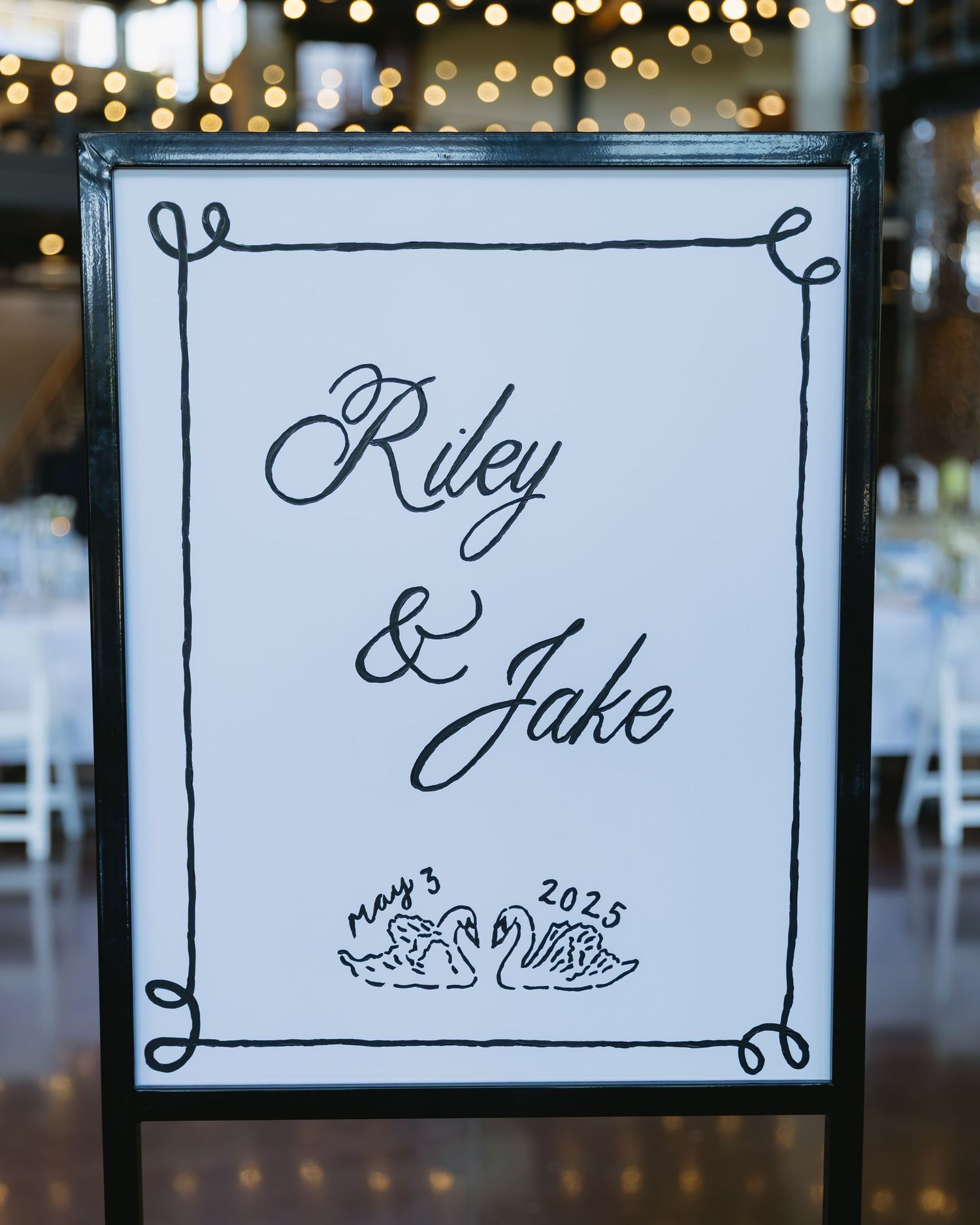 Elegant wedding sign showing names 'Riley & Jake' in cursive script with decorative swan motif in a black frame.