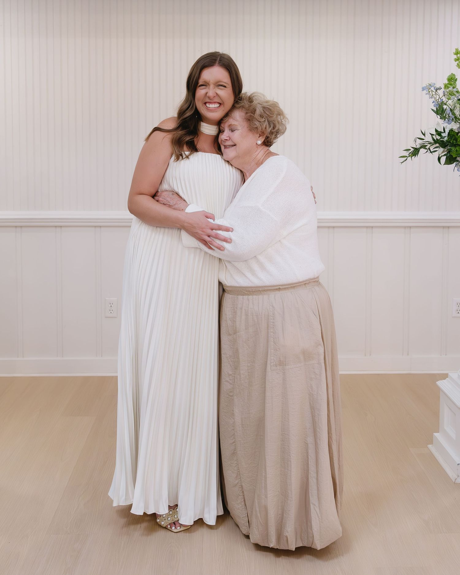 Two figures in white and beige outfits embracing and laughing together indoors.