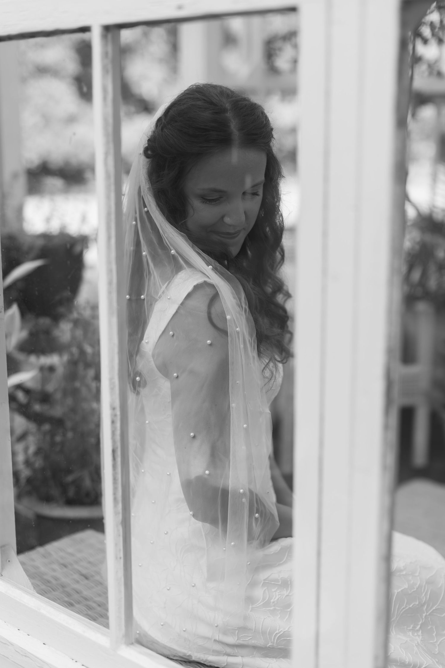 Black and white wedding photo showing a bride in white veil looking down through glass window panes.