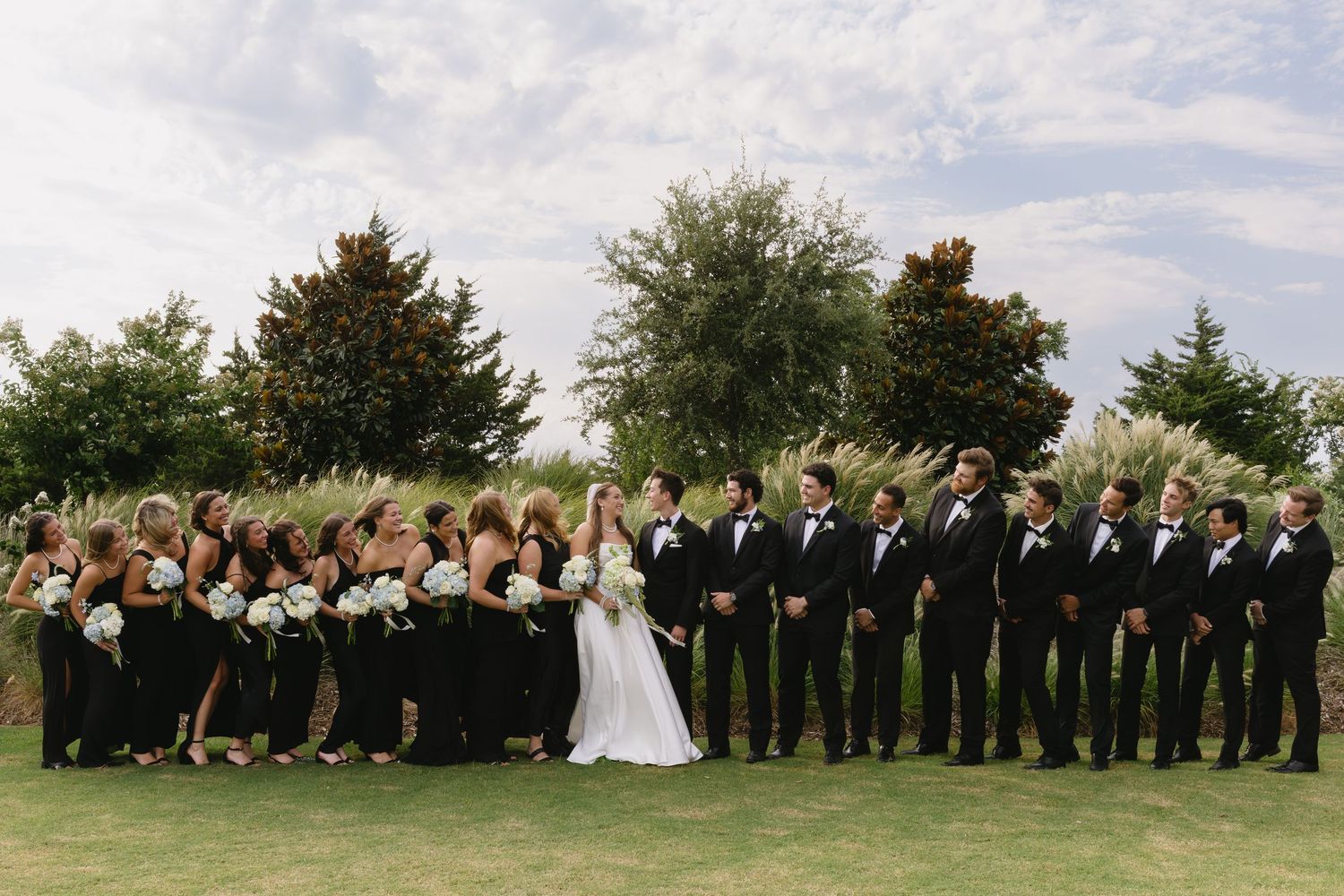 Large wedding party lined up outdoors with bridesmaids in black and groomsmen in tuxedos.