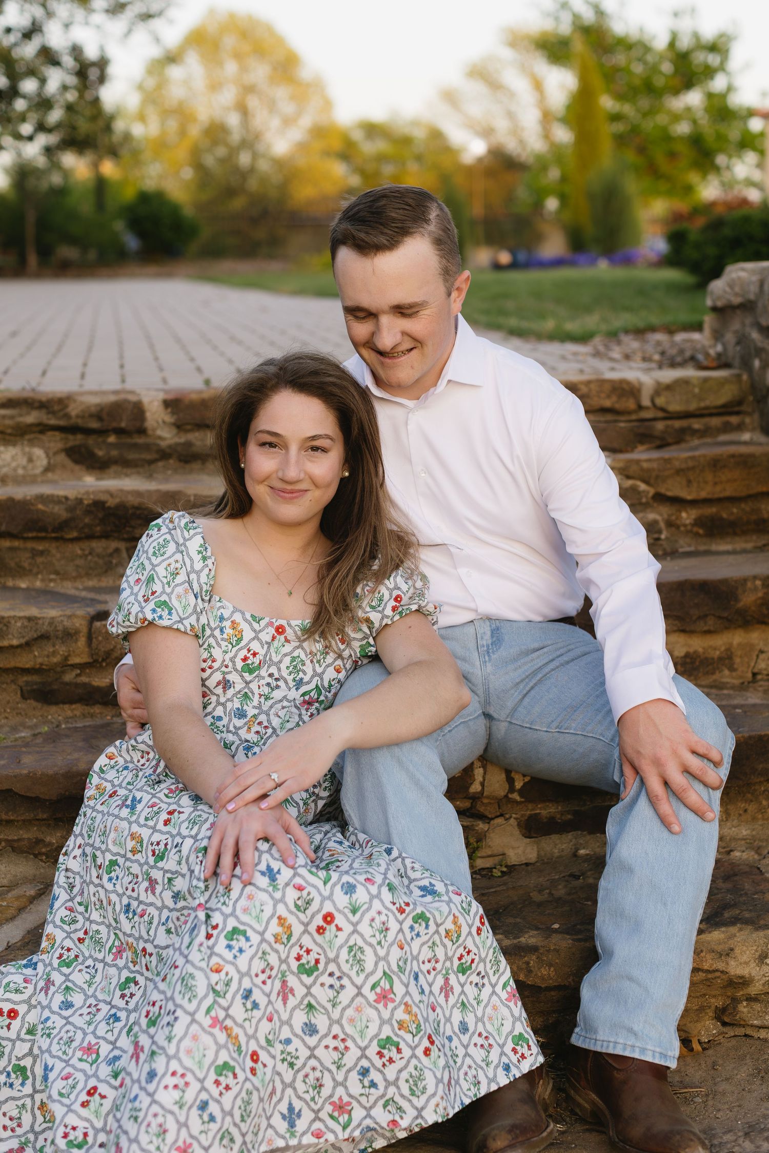 A couple sits together on stone steps surrounded by autumn foliage and trees.