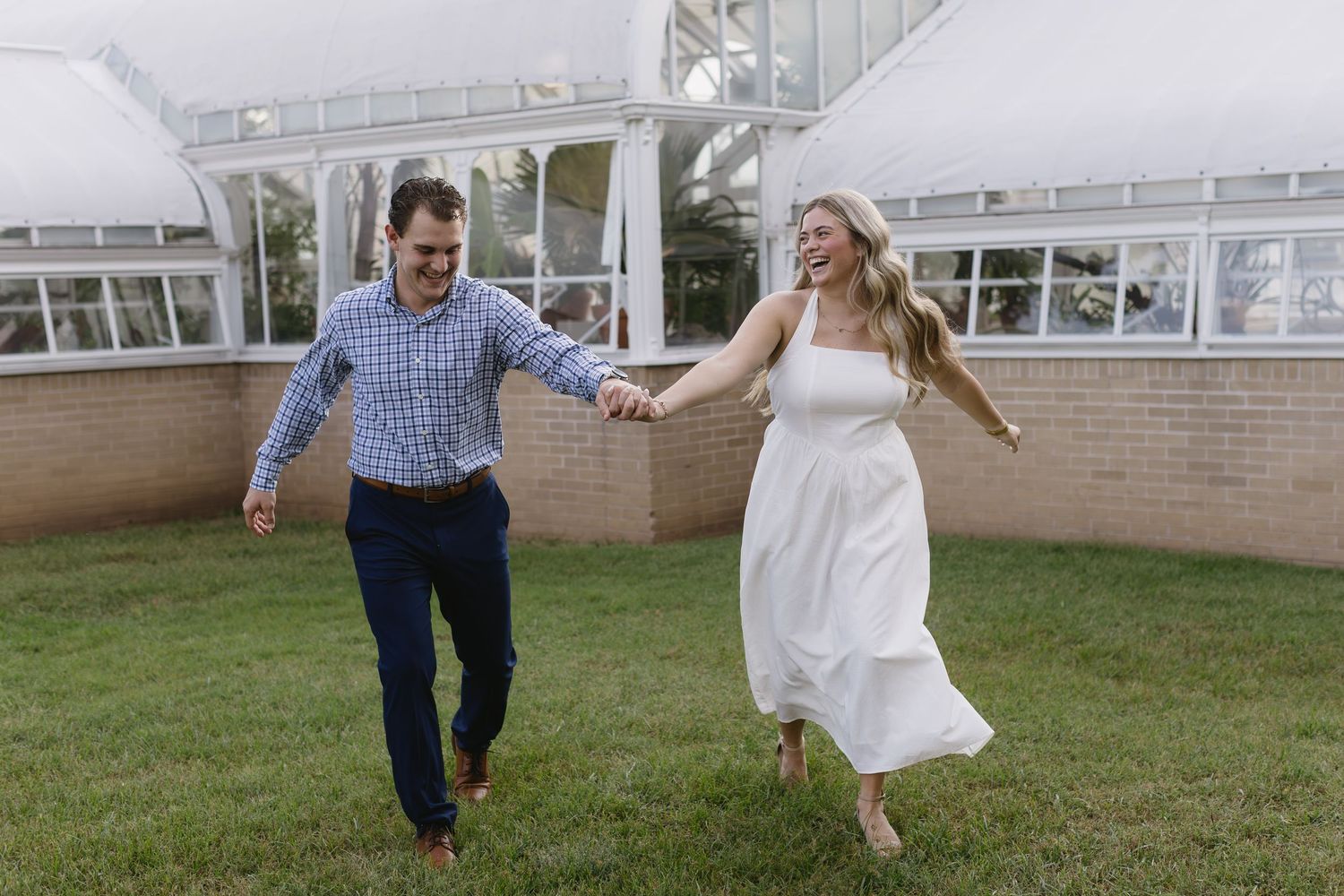 A couple in blue shirt and white dress holds hands while running together on a grassy lawn outdoors.