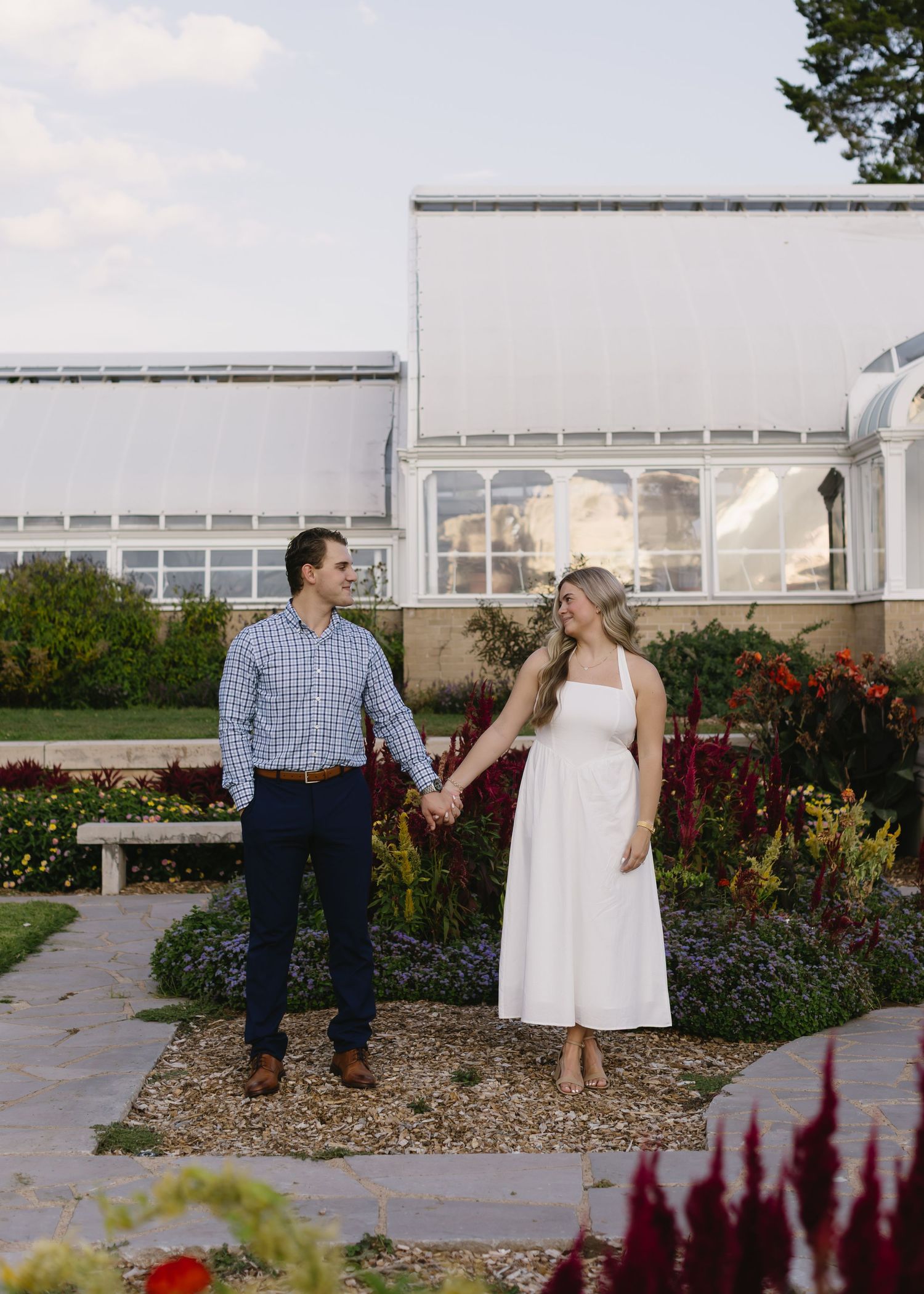 A couple shares a romantic moment in a garden with greenhouse structures and colorful flowers in the background.