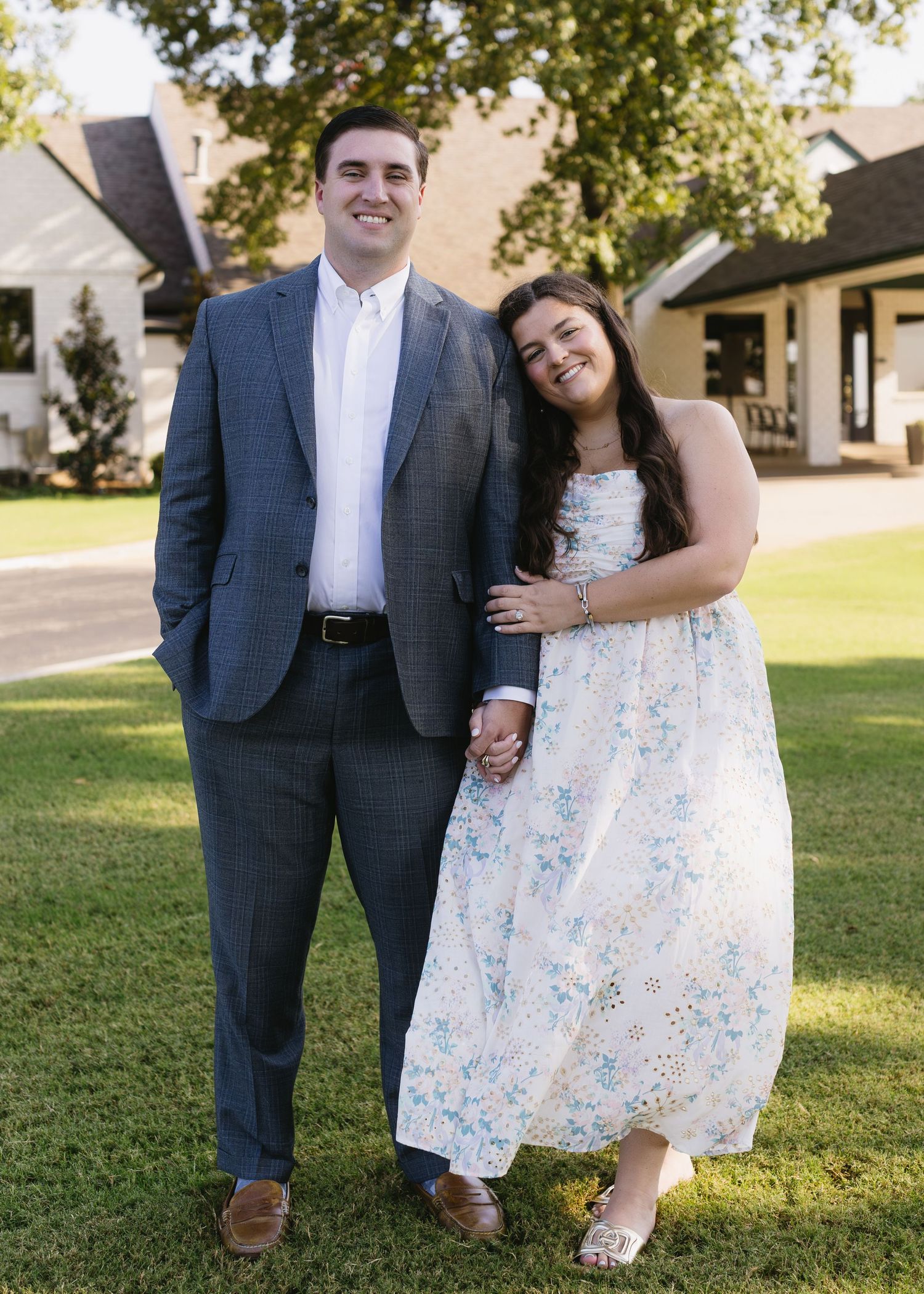 A couple in a floral dress and navy suit pose playfully together on a sunny lawn.