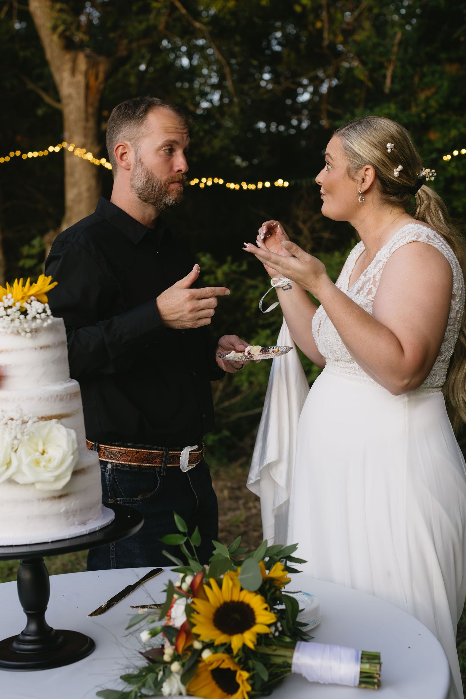 A couple shares their wedding cake during a reception decorated with sunflowers and string lights.