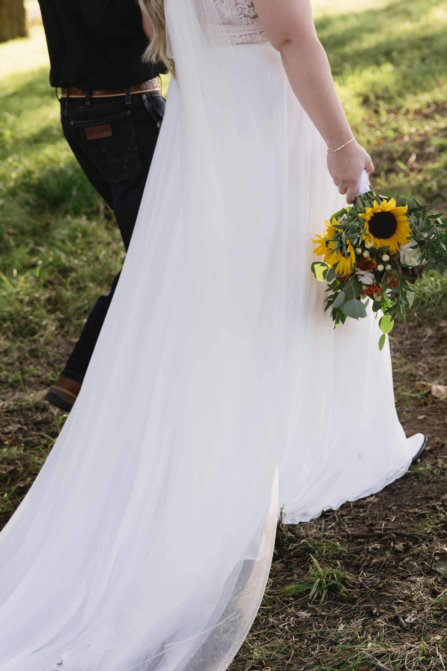 Close-up details of white wedding dress and sunflower bouquet against natural outdoor setting.