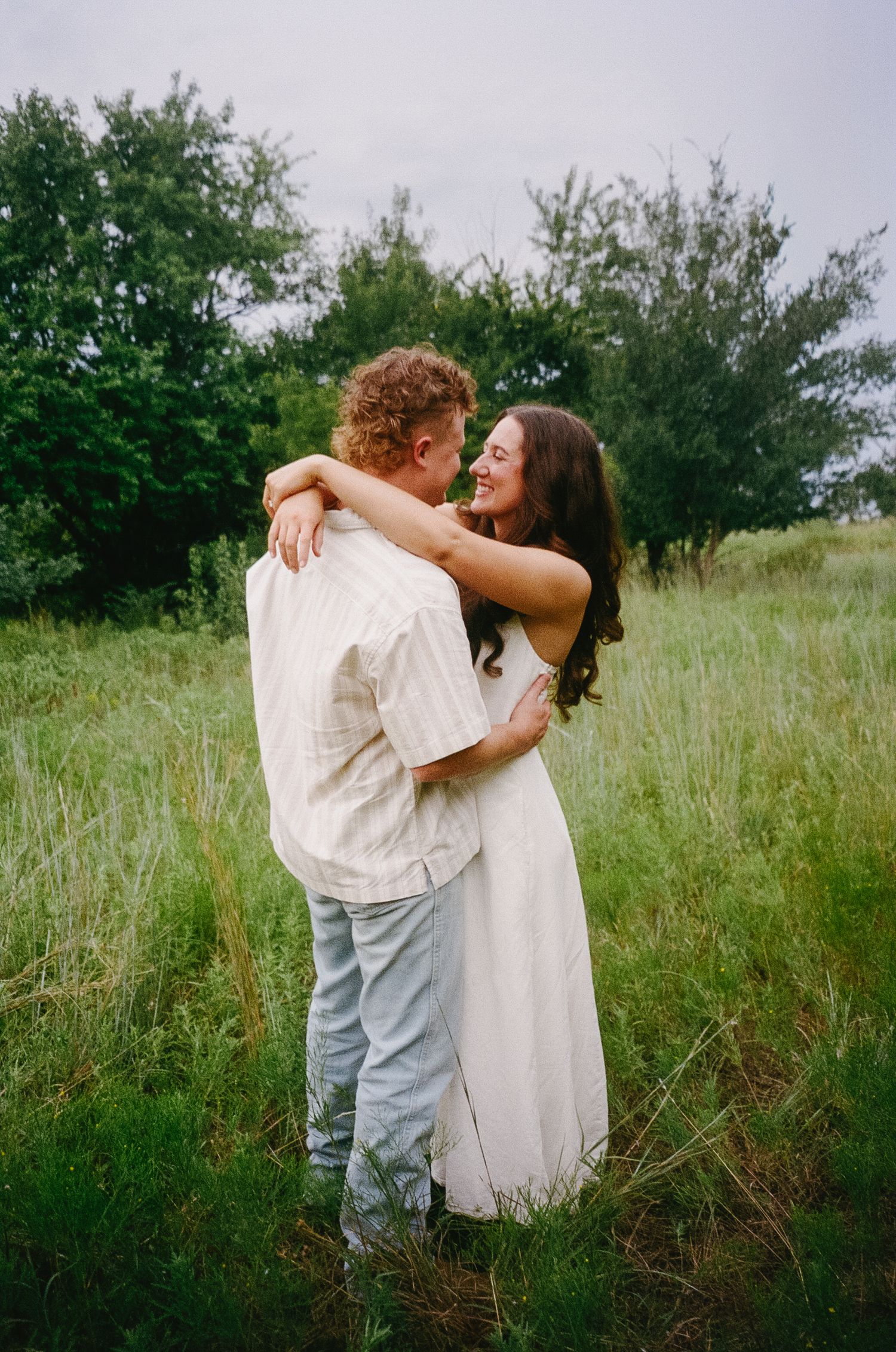 A romantic couple embraces in a sunlit meadow field against a backdrop of green trees and tall grass.