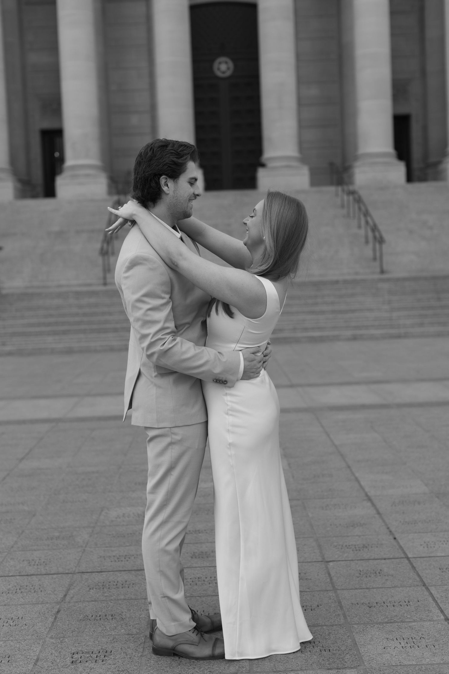A couple in coordinating beige and white outfits share a romantic moment on stone steps of a grand building.