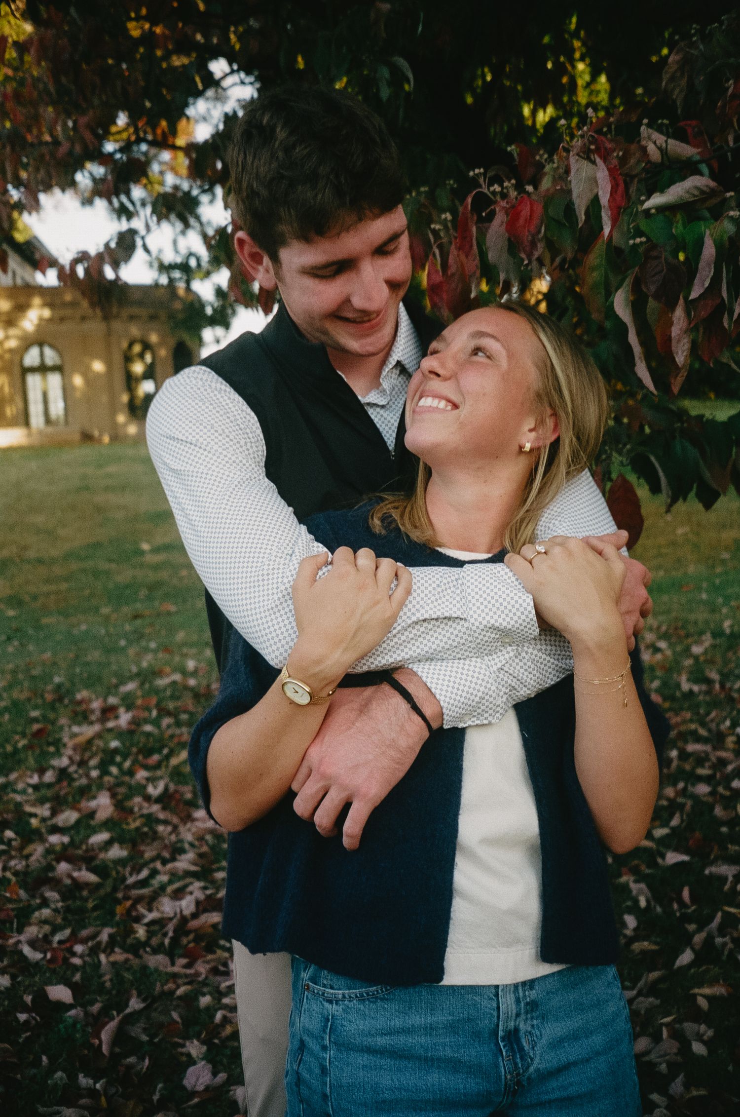 A couple embraces outdoors in autumn, with fallen leaves on the ground and trees in the background.