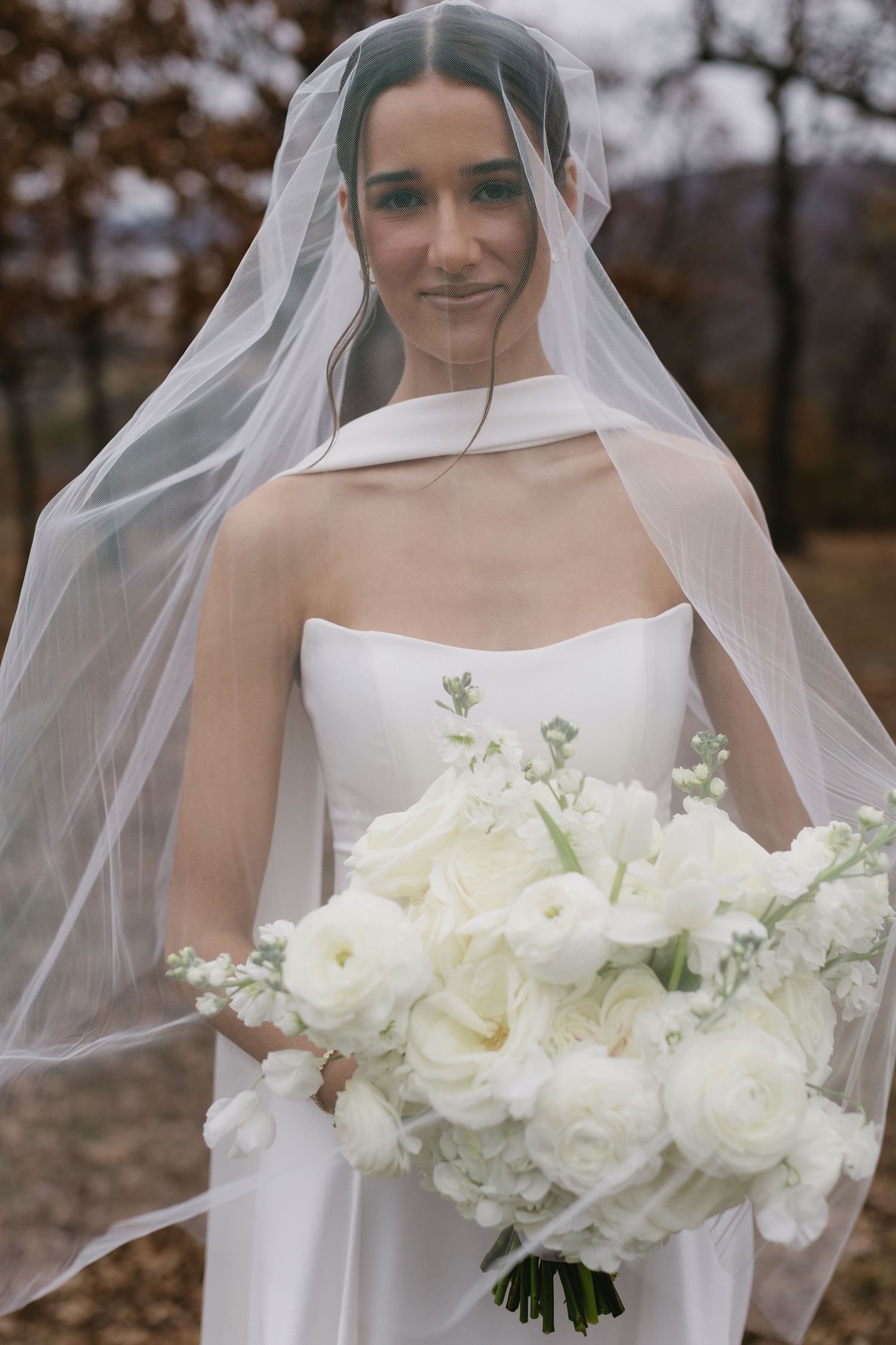 A bride in a strapless white gown holds a bouquet of white flowers under her veil.
