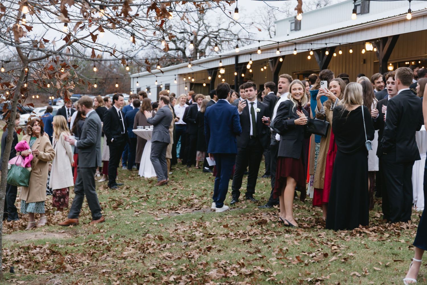 Large group of wedding guests gather outdoors under string lights and autumn trees.