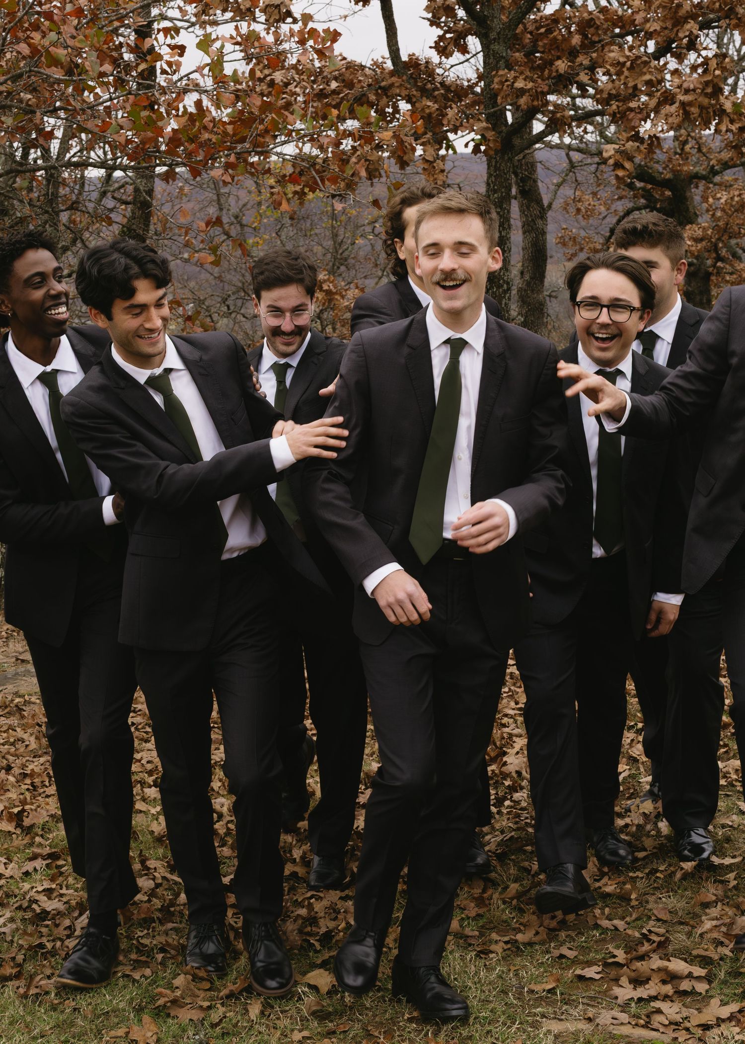 A group in formal black suits share laughter during an outdoor autumn wedding photoshoot.