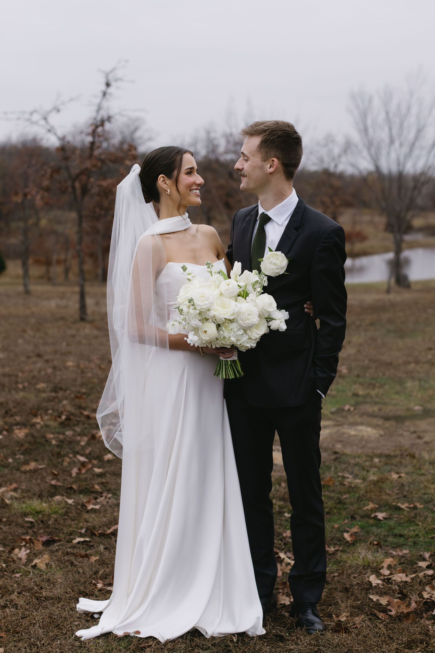 Newlyweds share an intimate moment outdoors with white bouquet against autumn landscape.