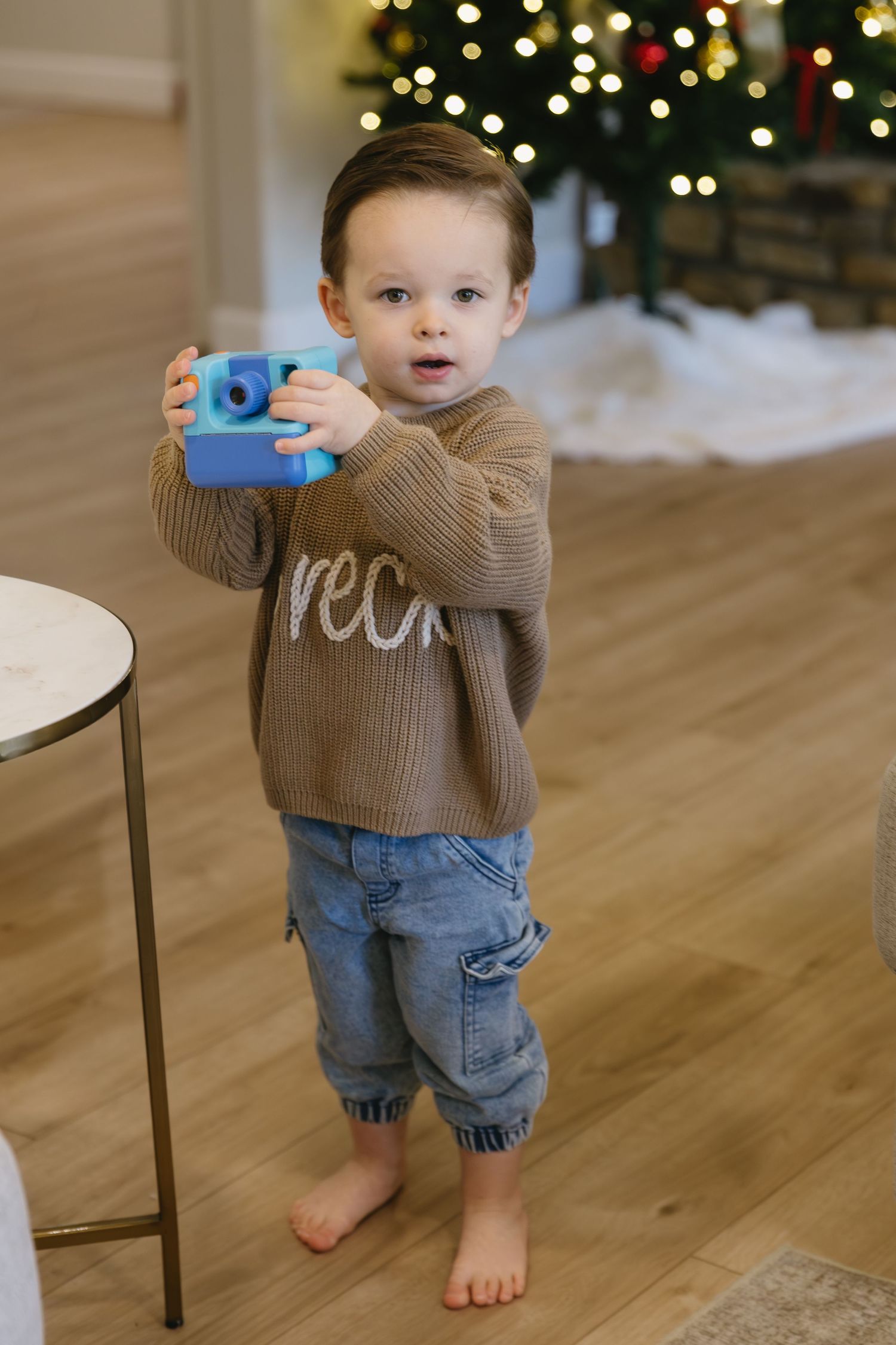 Toddler in brown sweater and jeans standing barefoot near Christmas tree lights.