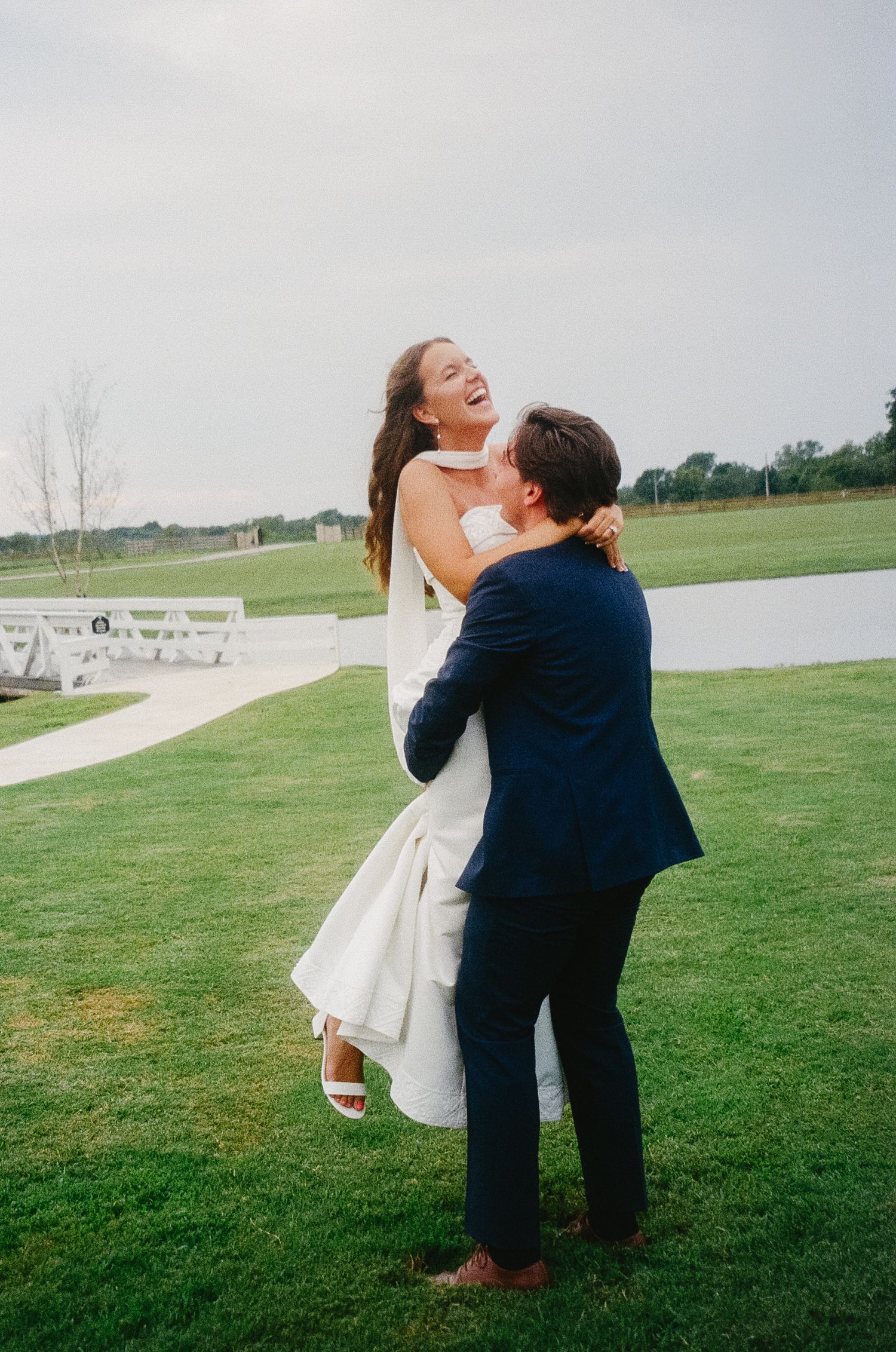 A couple in wedding attire joyfully embrace and dance together on a green lawn under overcast skies.