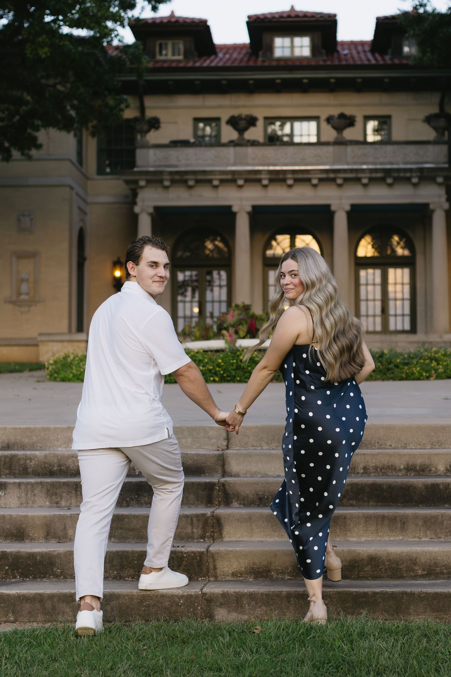 A couple poses playfully on steps of an elegant mansion, coordinating in polka dots and white.