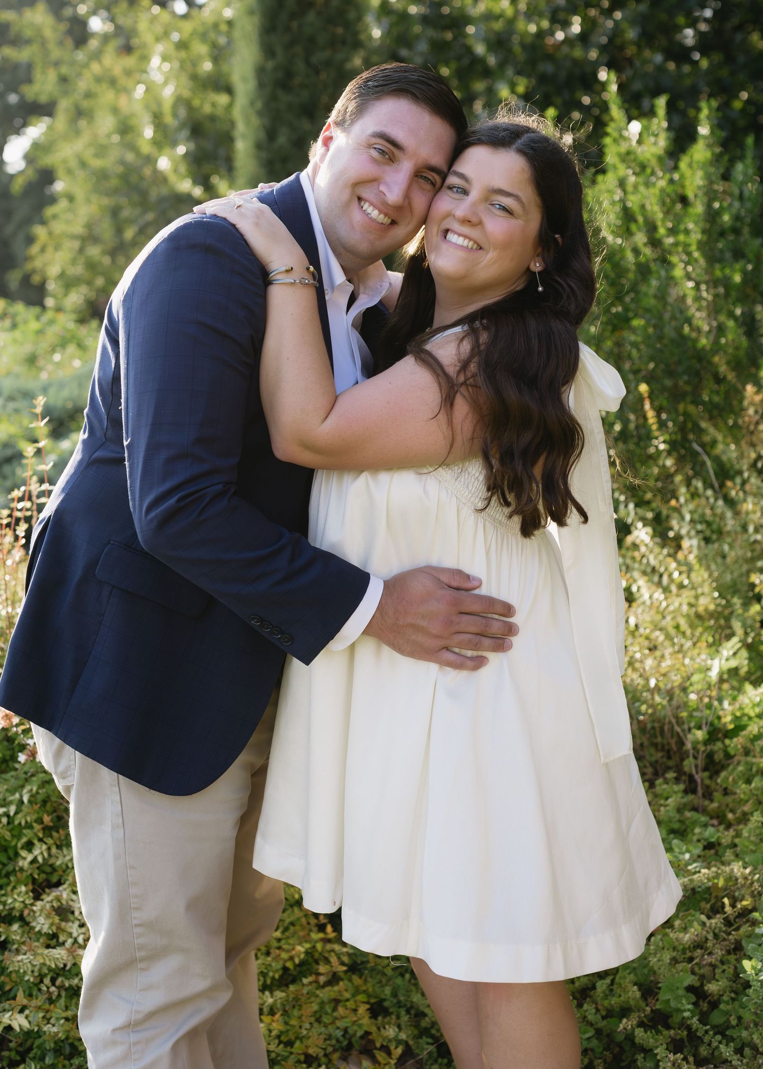A couple embraces and smiles while standing in front of green foliage.