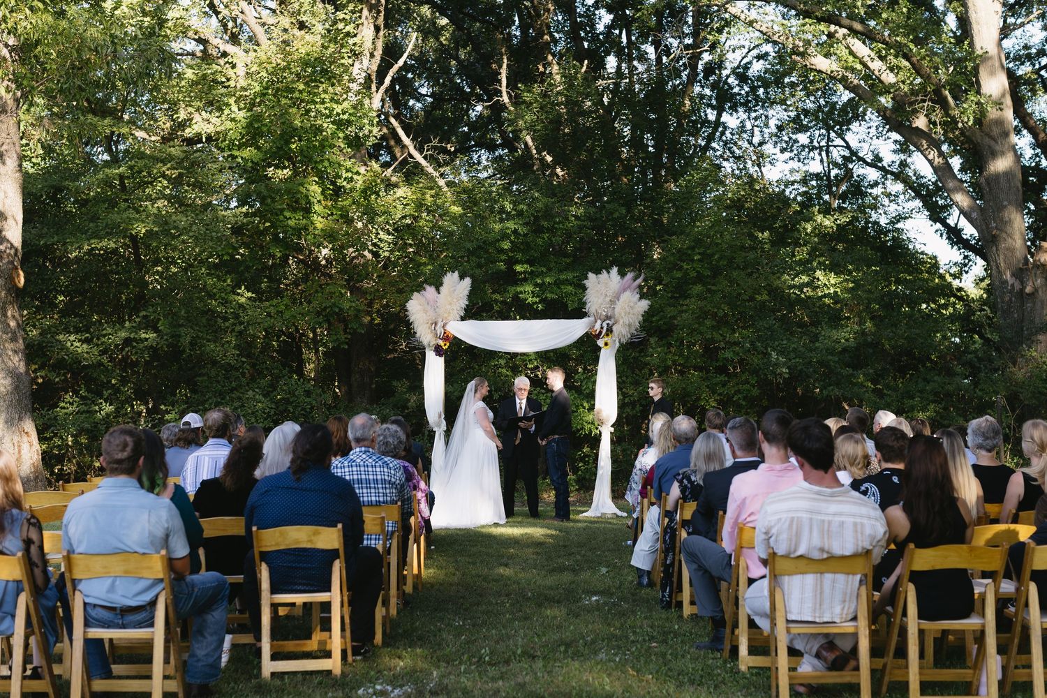 Outdoor wedding ceremony setup with wooden chairs arranged under tall trees.