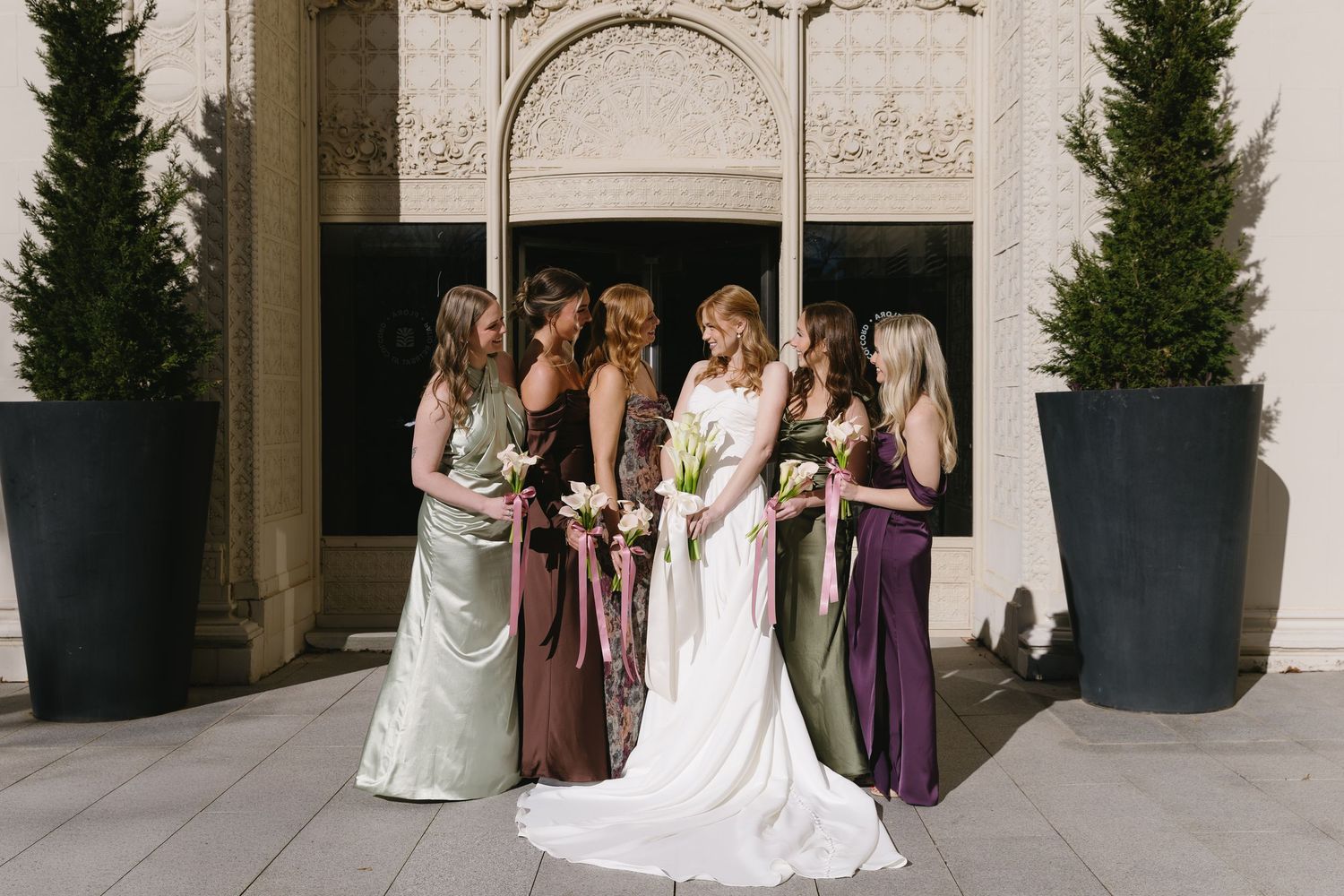 Wedding party poses elegantly outside venue with bridesmaids in varying shades of purple and mint dresses.