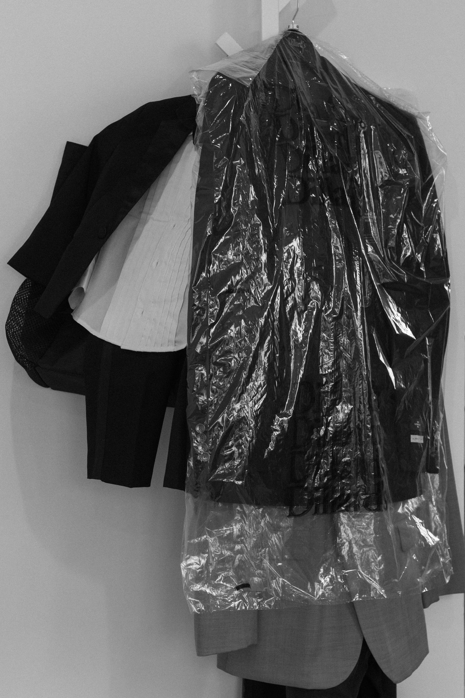 Dry cleaned clothes in plastic garment bag hanging on white wall with black coat visible.