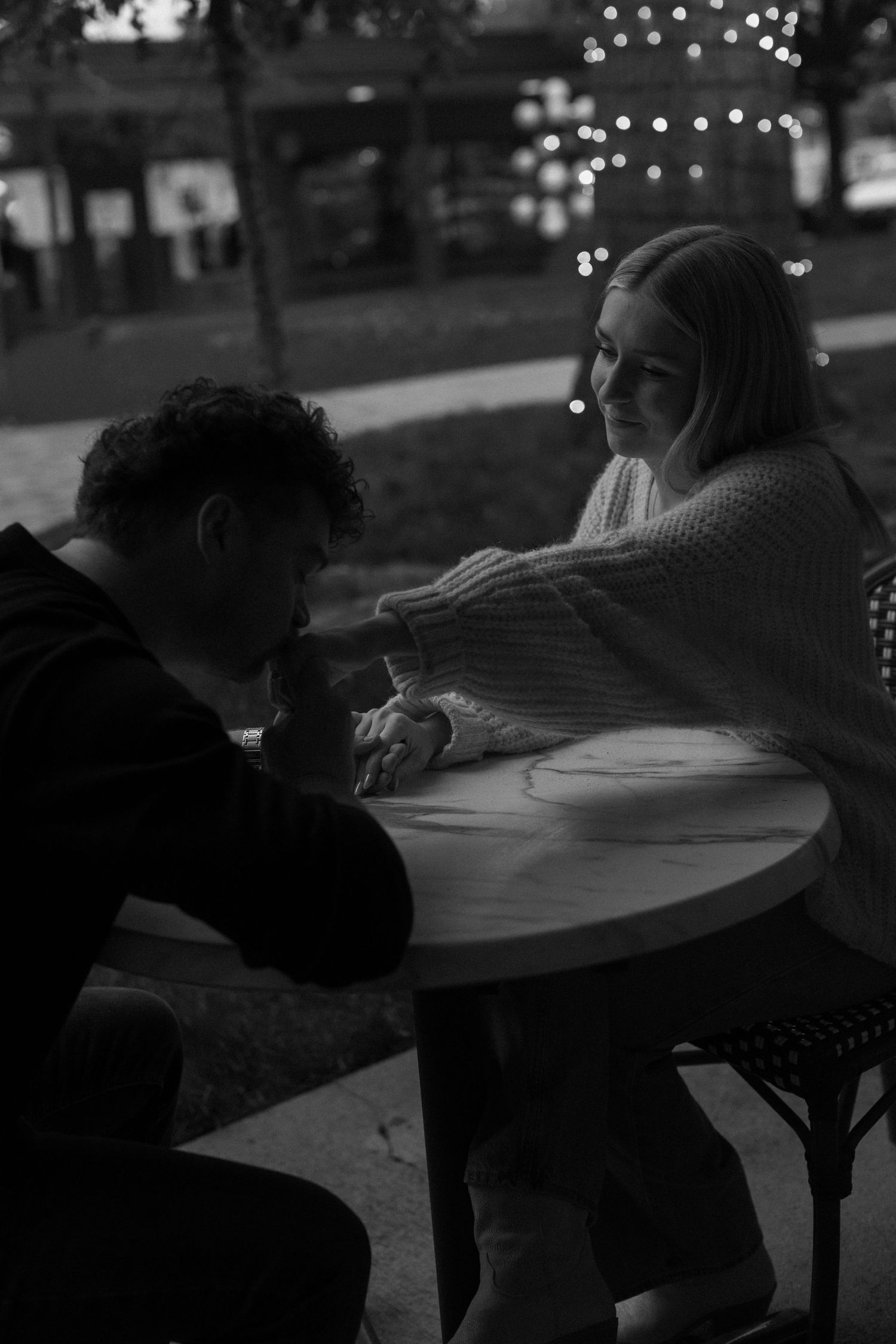 A romantic black and white photo of a couple sitting at a table outdoors with twinkling lights in the background.