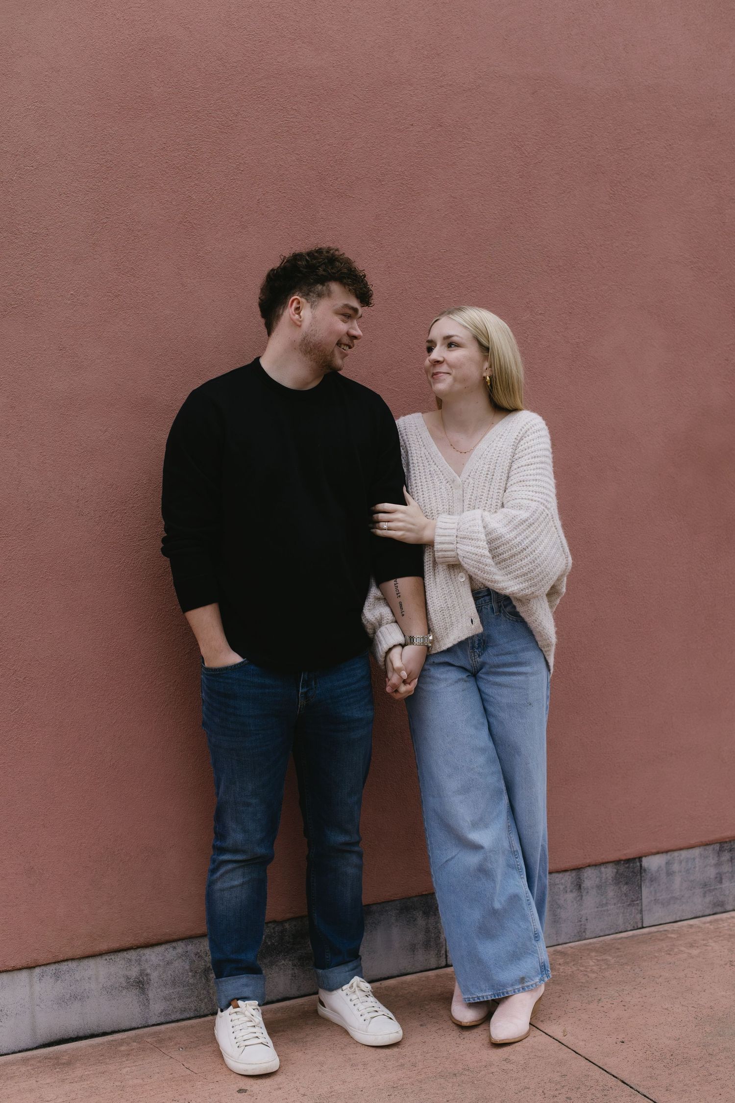 Couple in casual attire poses playfully against a pink wall in a series of candid shots.
