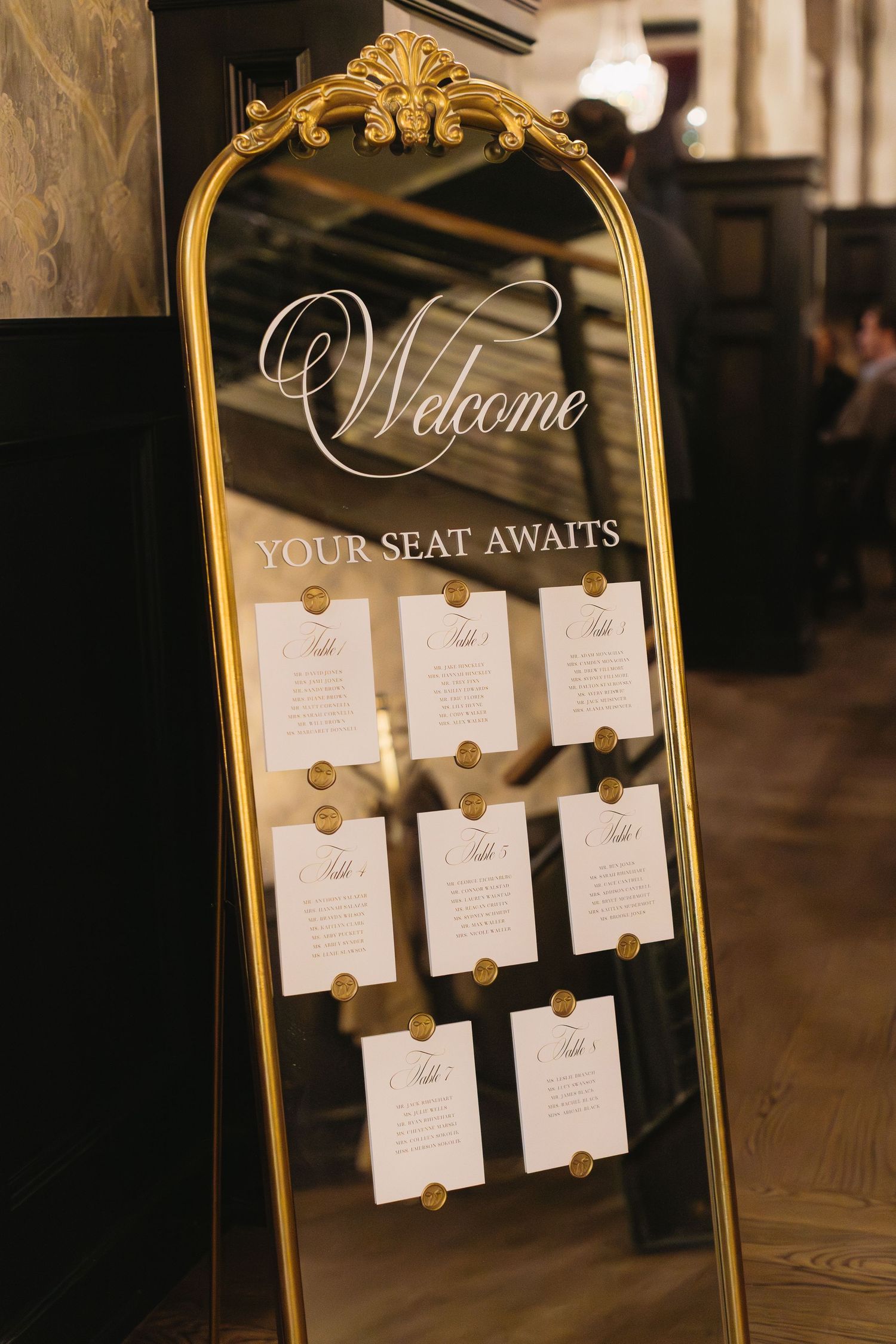 Elegant gold-framed mirror displays wedding seating assignments with 'Welcome' text and white cards arranged neatly.